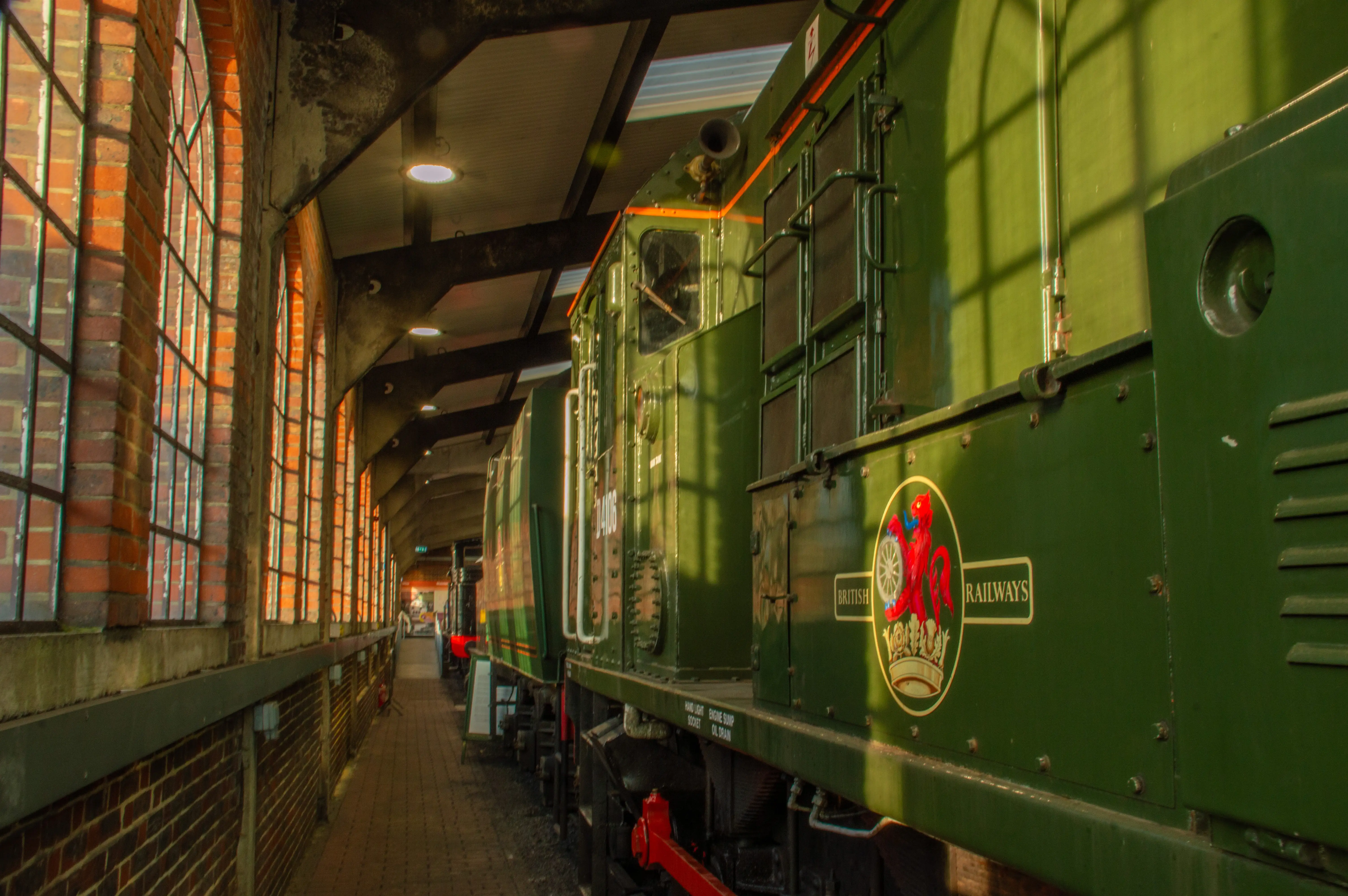 Side view of a dark green vintage British Railways steam locomotive at a station platform, with sunlight casting shadows from arched brick windows onto the walkway.