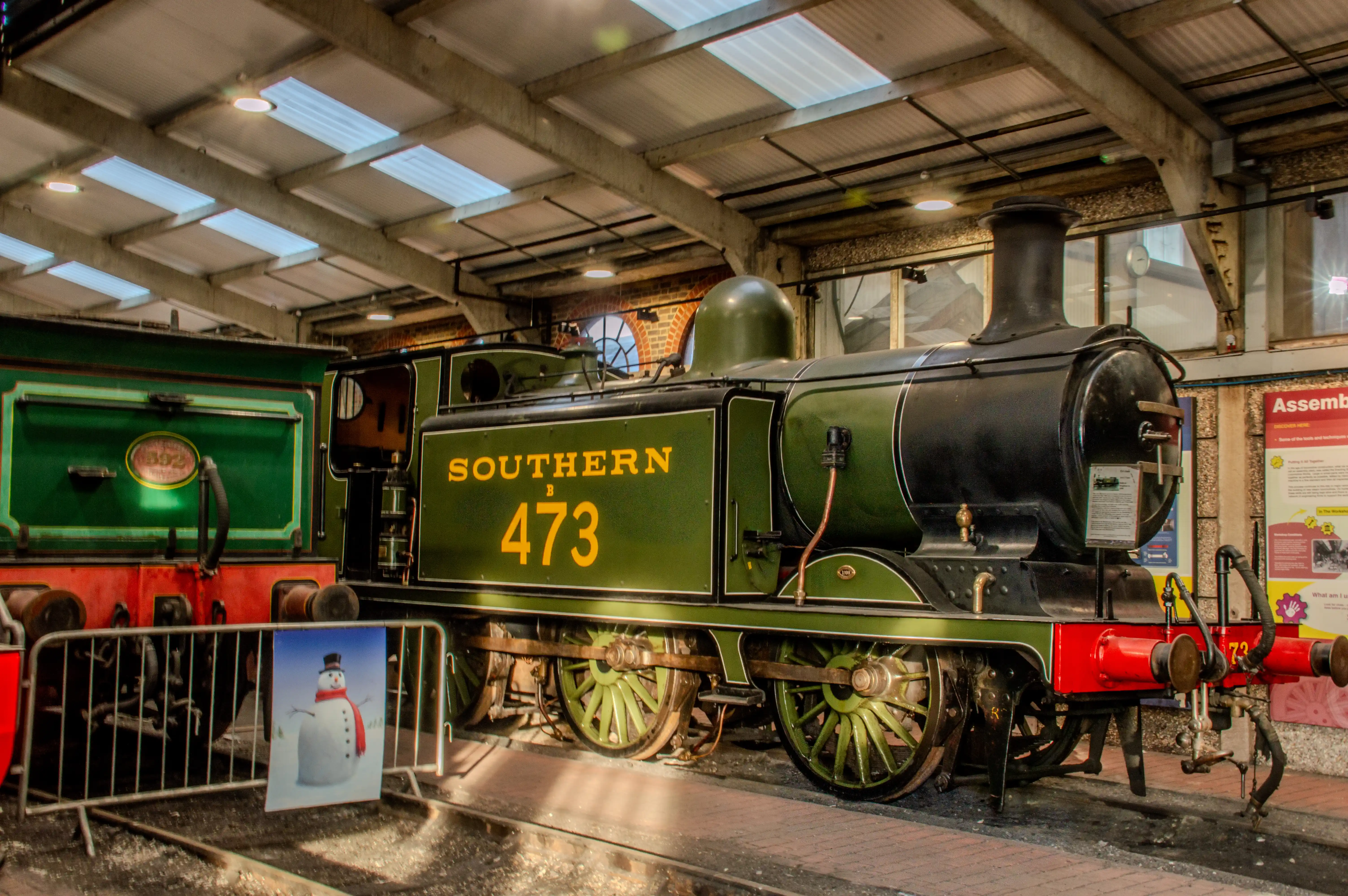 Green and black steam locomotive numbered 473 with &laquo;Southern&raquo;, written in yellow, parked indoors with skylights in the ceiling. A snowman sign is visible in front of a barrier.