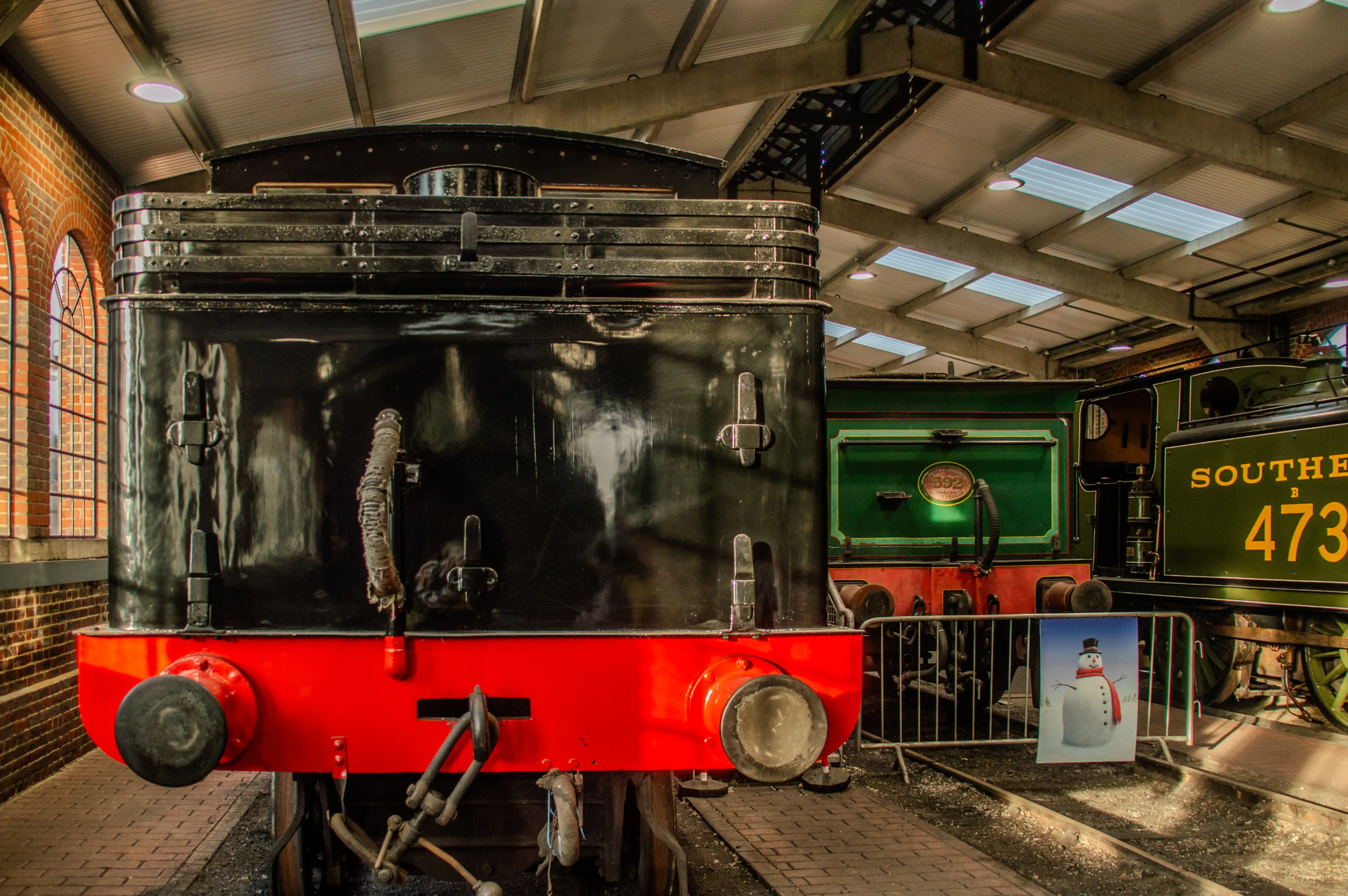 The front of a black and red steam locomotive is prominently featured, with other vintage trains visible in the background within a museum setting.