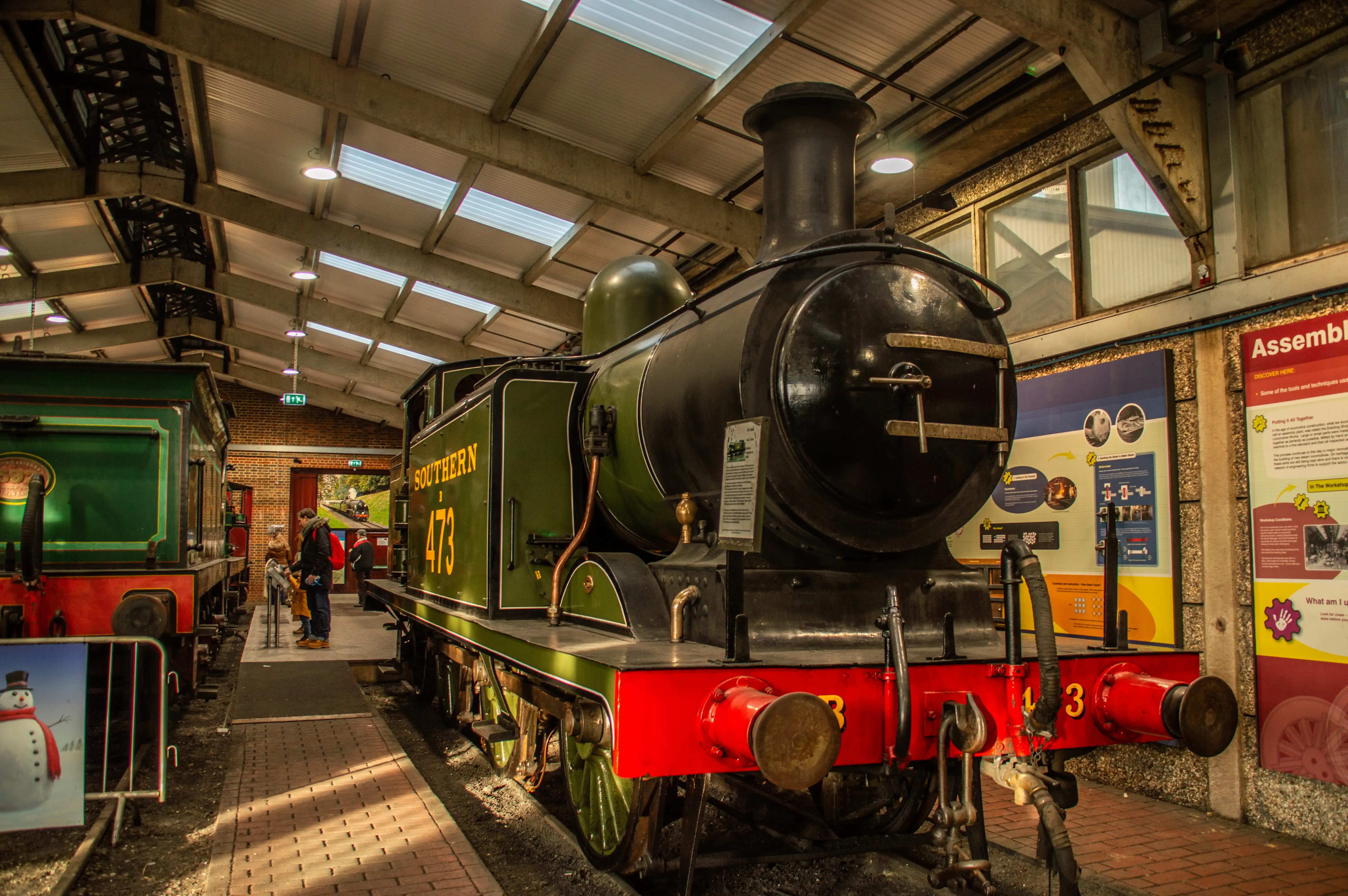 Black steam locomotive numbered 75027 with a red buffer beam inside a railway shed, flanked by other engines and visitors on a brick walkway.