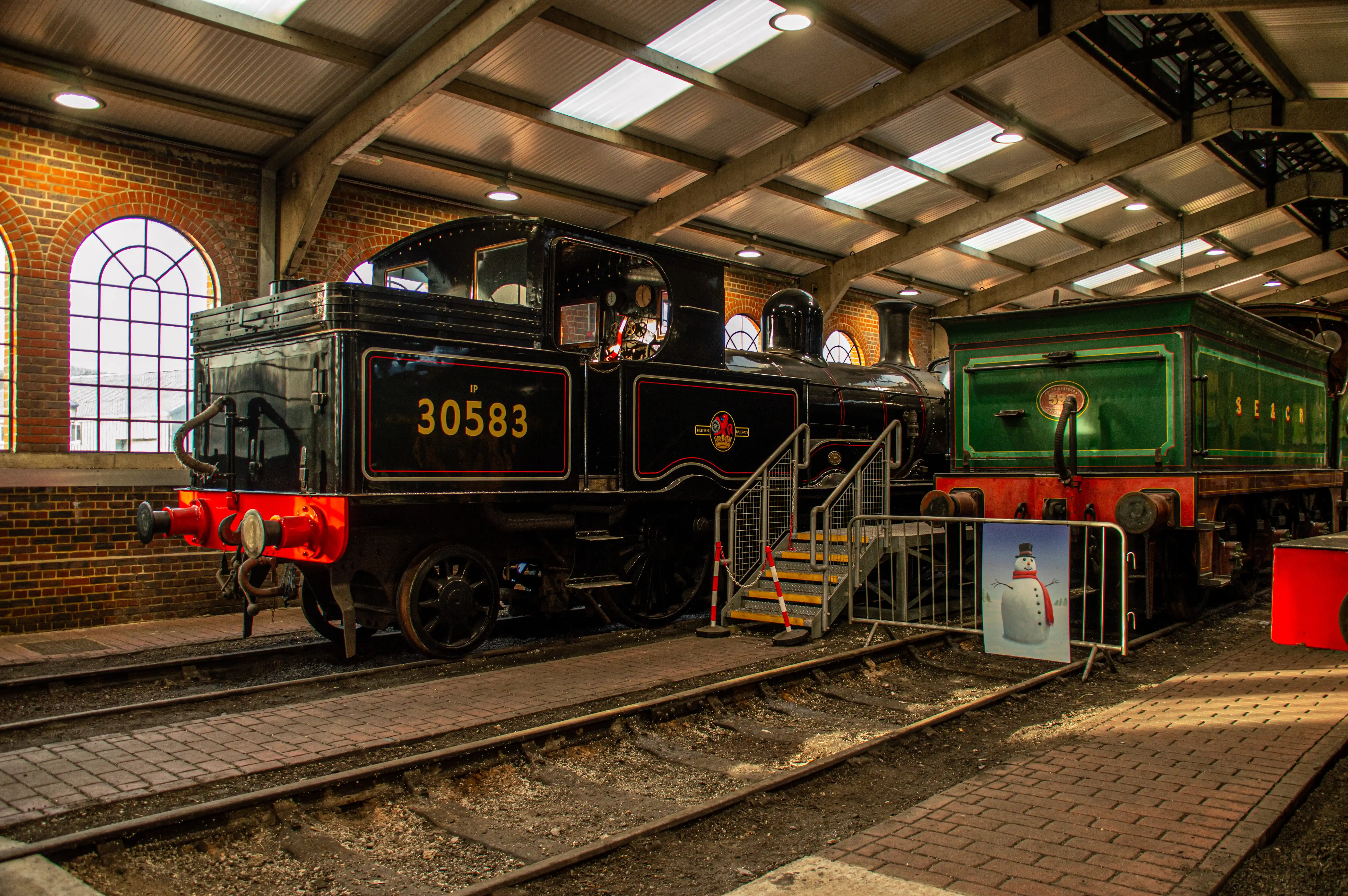 A black steam locomotive with the number 30583 is parked on railway tracks inside a brick building with a corrugated roof and arched windows. A green railway carriage is parked next to it, and a poster of a snowman with a red scarf is visible.