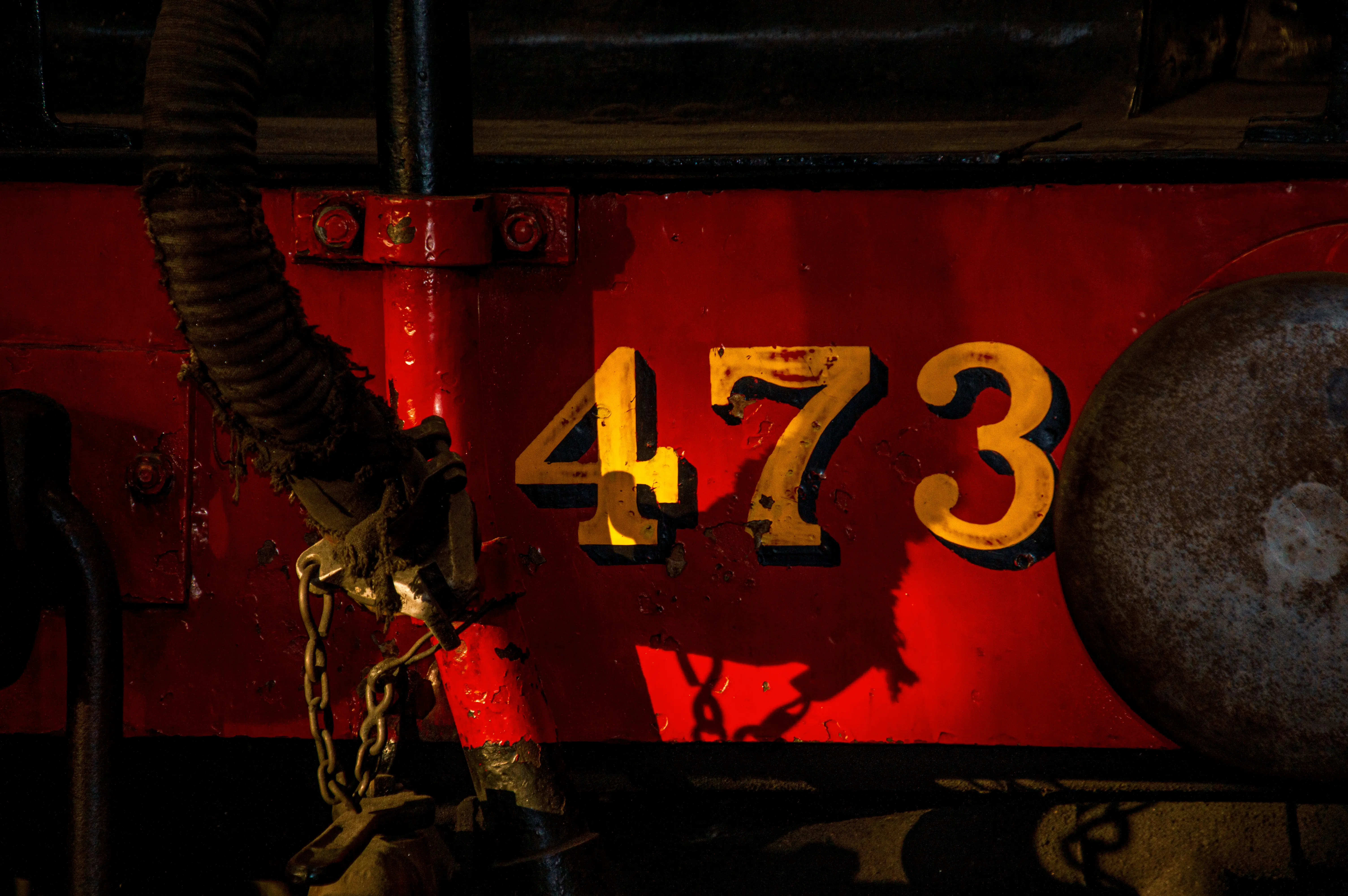 Close-up of the number 473 in yellow with a dark outline painted on a dark red train carriage. Mechanical chains and parts are visible on the left.