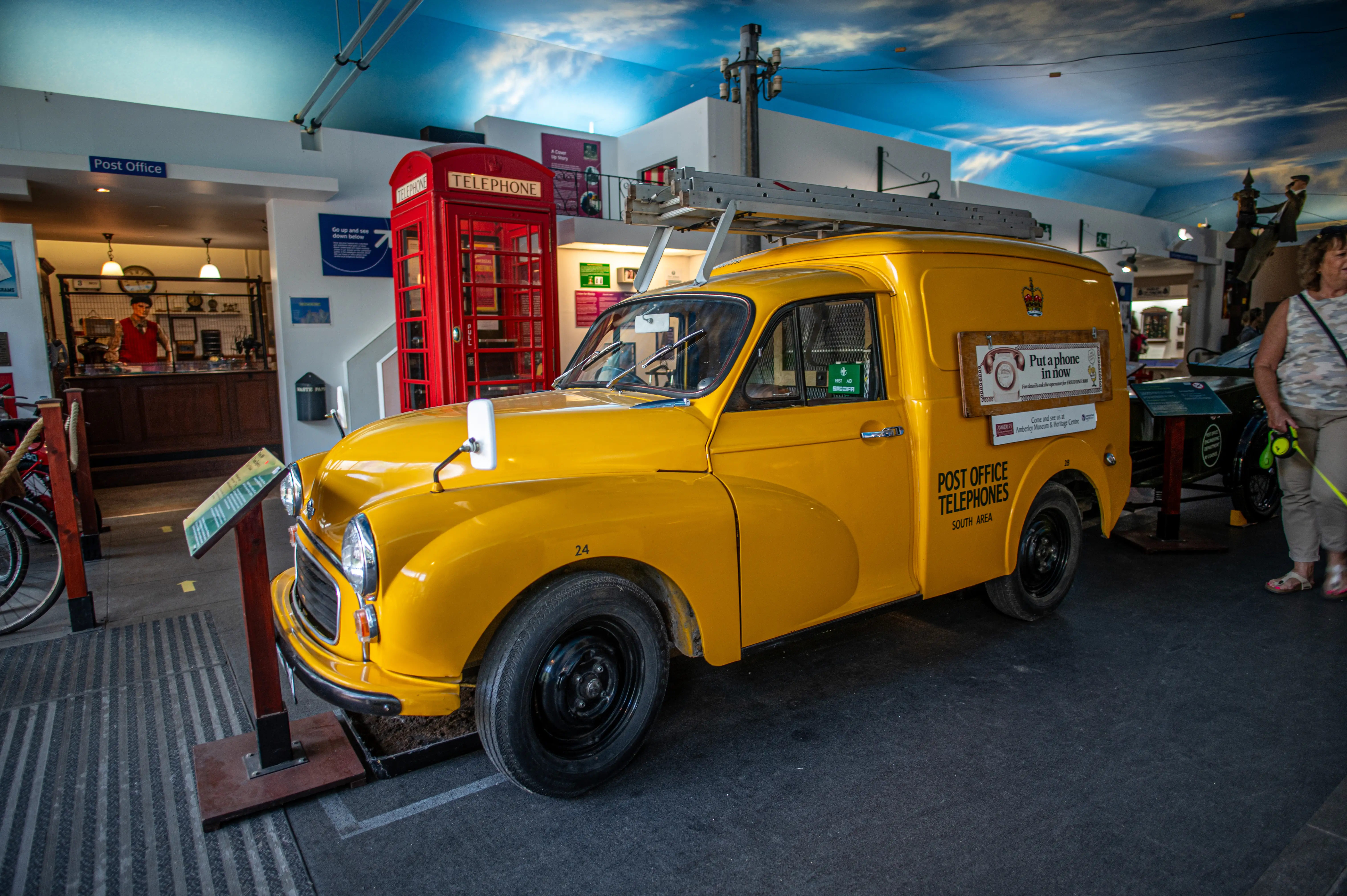 A bright yellow vintage Post Office van is on display next to a red telephone box inside a museum. The van has POST OFFICE TELEPHONES written on its side. A woman and her dog are in the background.