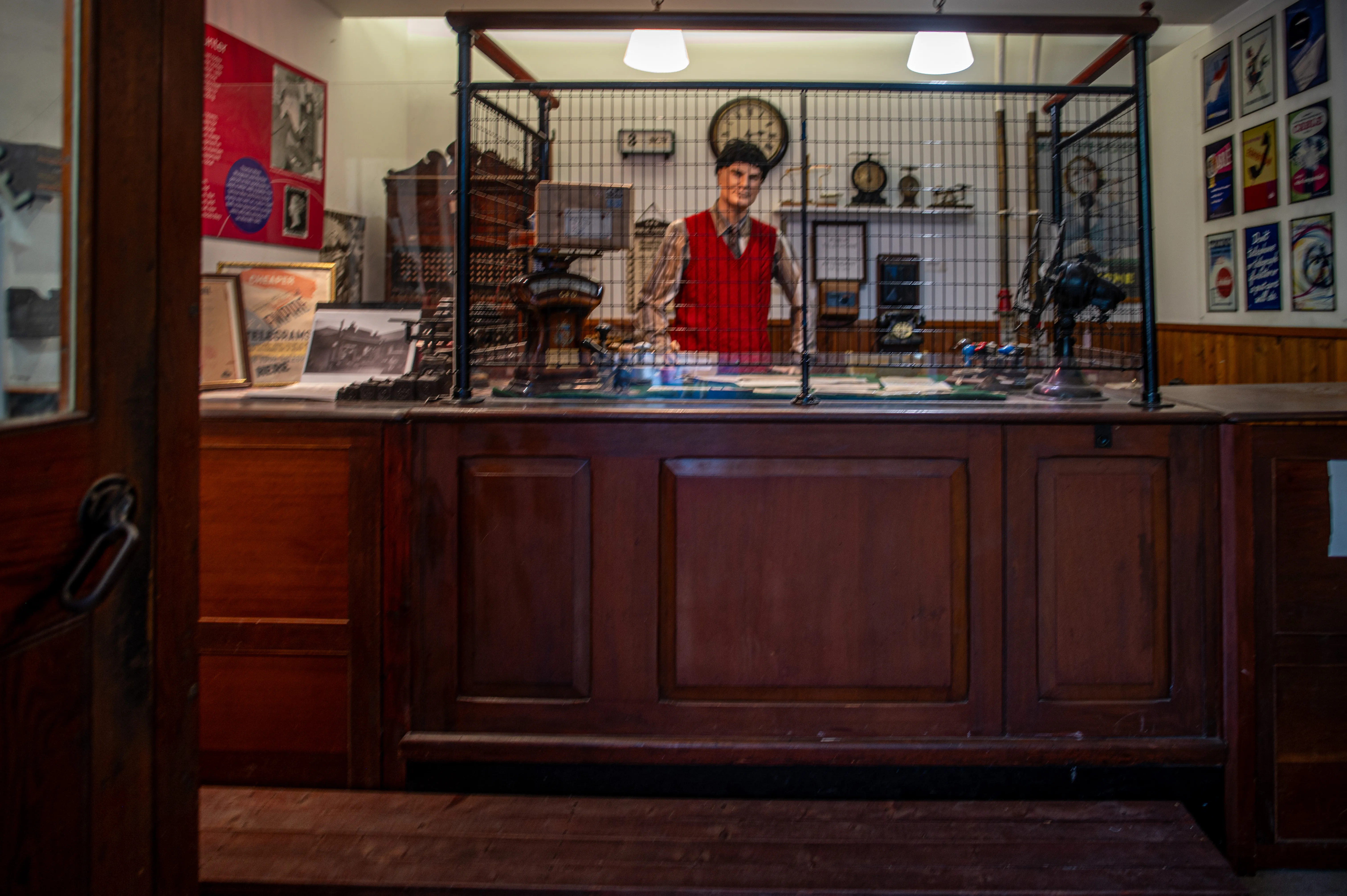 A vintage shop counter made of dark wood, with a lifelike figure of a shopkeeper in a red vest and cap. The counter is enclosed by a metal grid, and the background features framed photographs, posters, and a clock