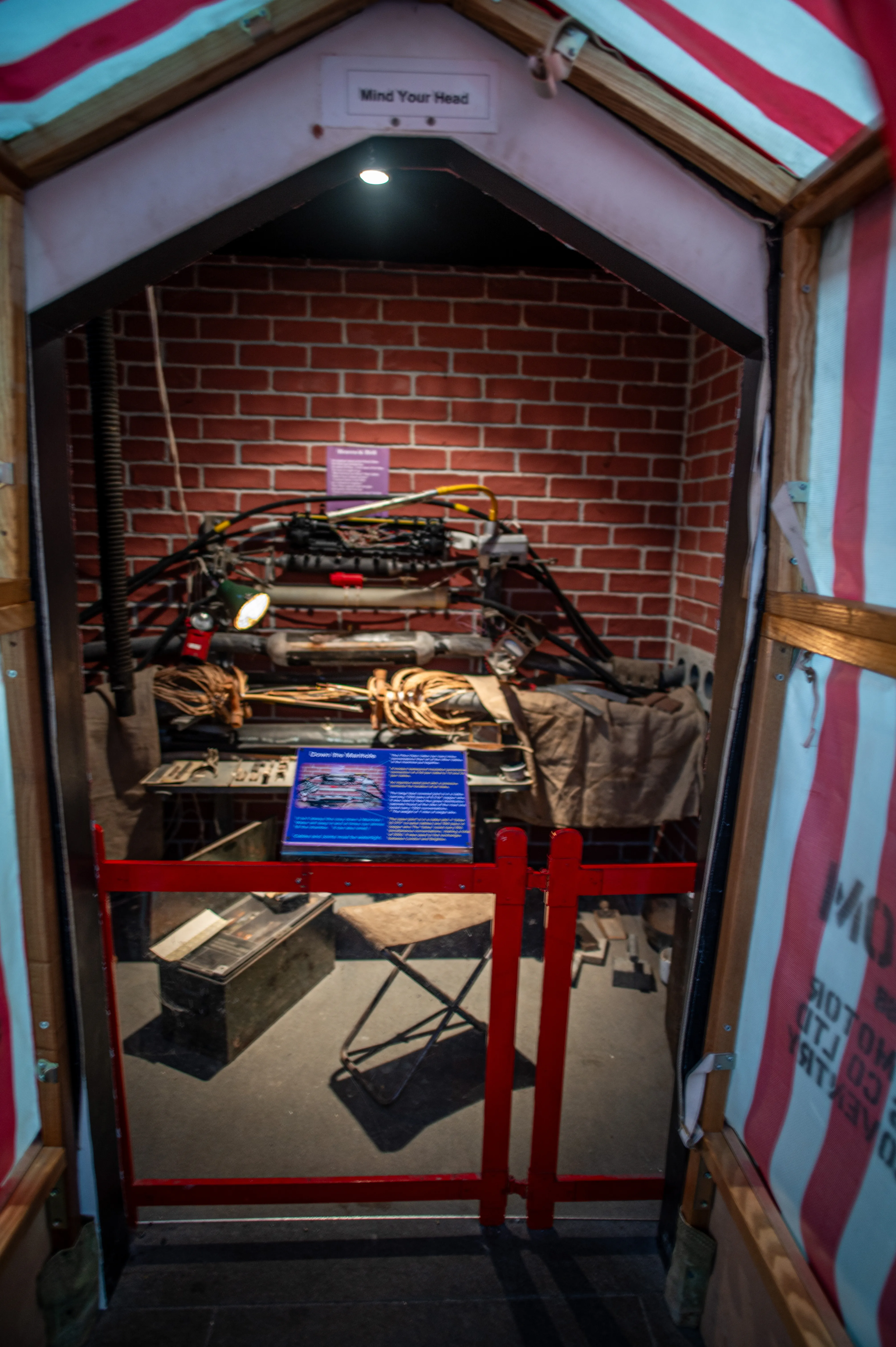  Inside a narrow exhibit recreating a Victorian sewer maintenance area, a complex array of pipes, wires, and tools is visible against a brick wall. A blue information plaque is in the foreground, and a red barrier partially obscures the view. A sign above the entrance reads &laquo;Mind Your Head.&raquo;