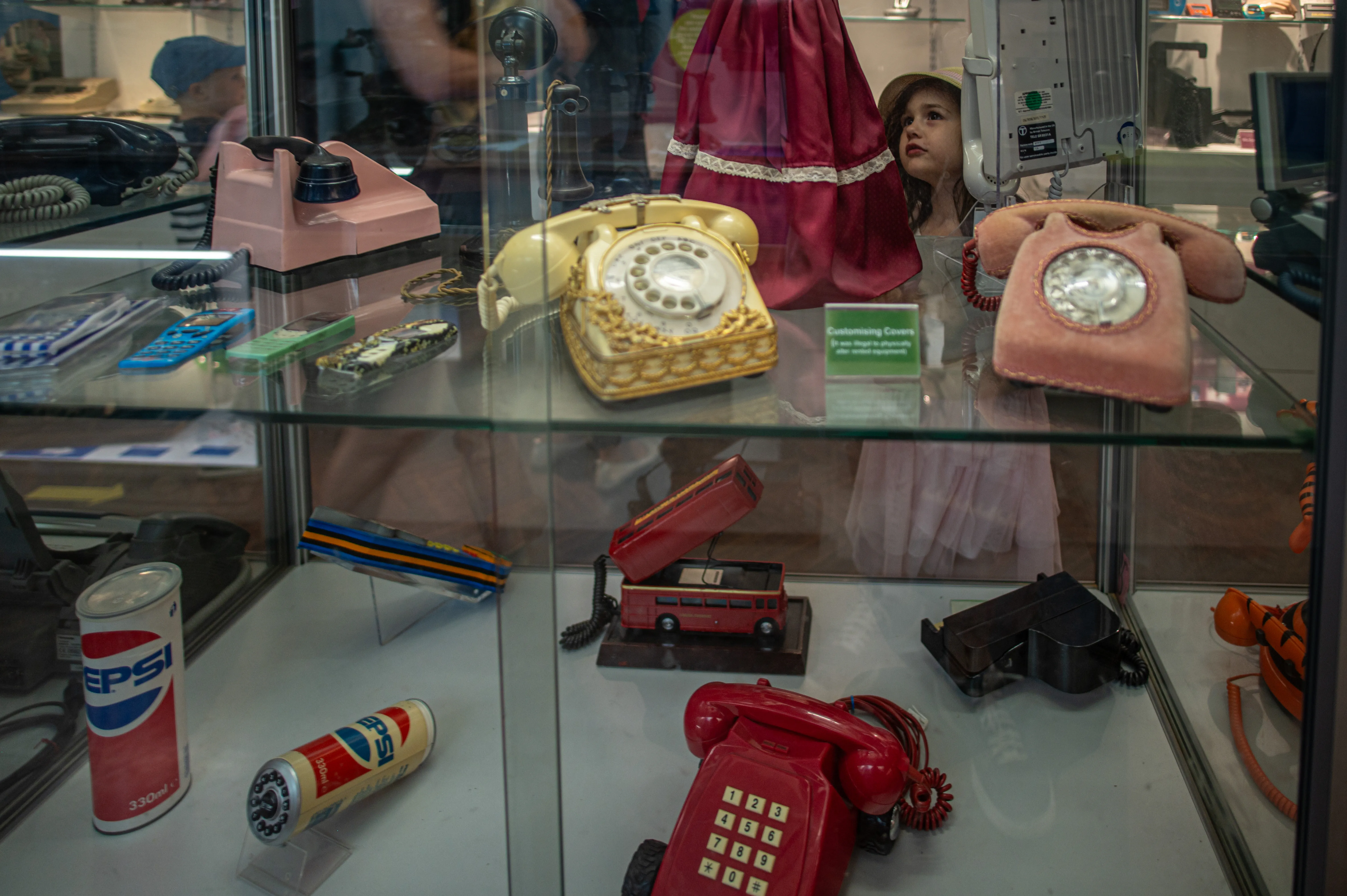 A glass display case with vintage telephones, including a pink rotary phone, a gold-toned decorative phone, and a red push-button phone. Other items include a Pepsi can, a small red double-decker bus model, and a 'Customising Covers' sign.