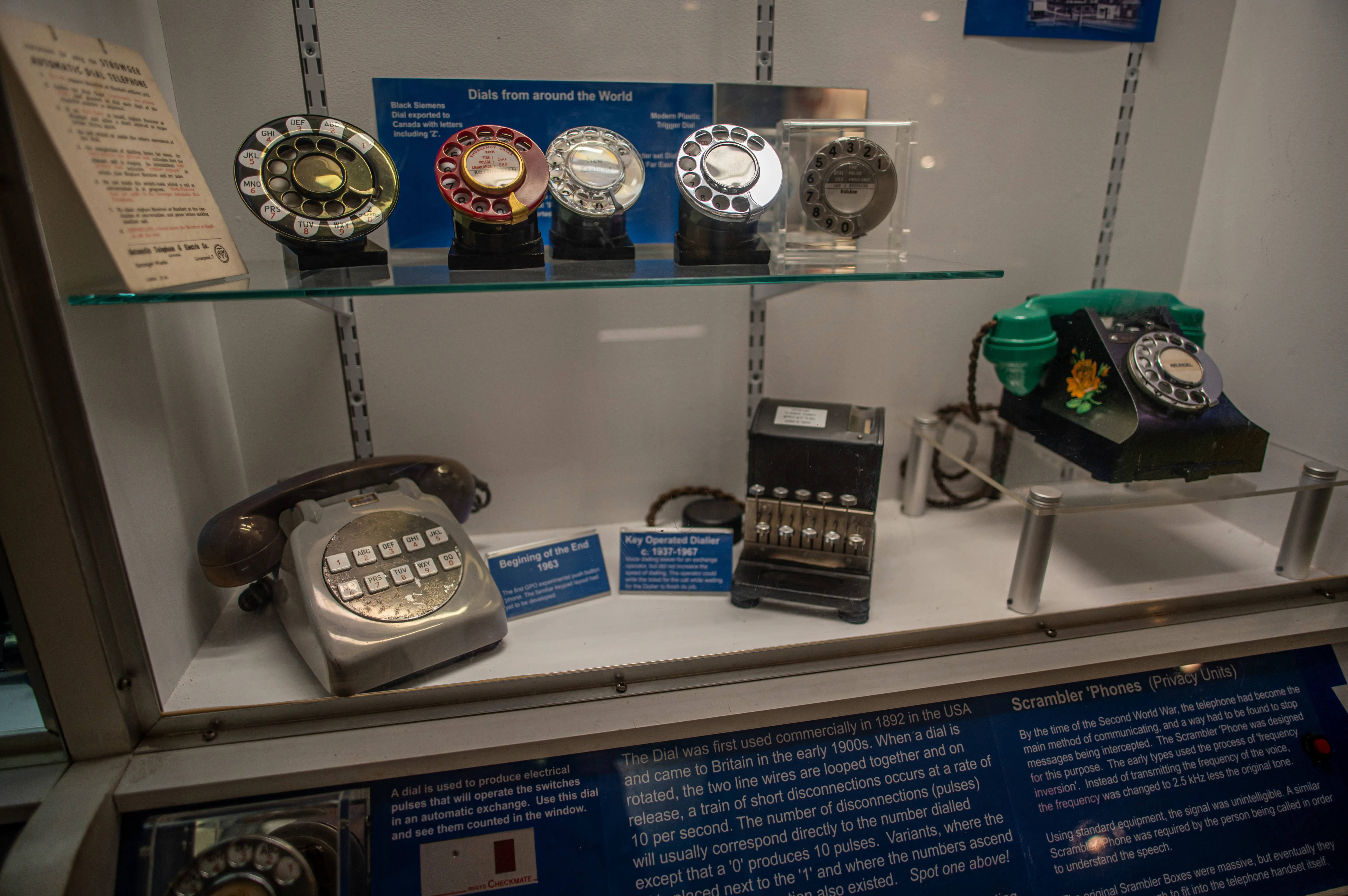 A museum display case containing vintage telephones and dial mechanisms. On the top shelf are several rotary telephone dials from different eras and countries, some made of metal and others of plastic. Below, on the middle shelf, sits a grey rotary dial telephone from 1963, a black key-operated dialer, and a decorative green telephone with a floral design. The bottom of the case features informational plaques detailing the history and function of the displayed items, including the evolution of the telephone dial and the use of scrambler phones.