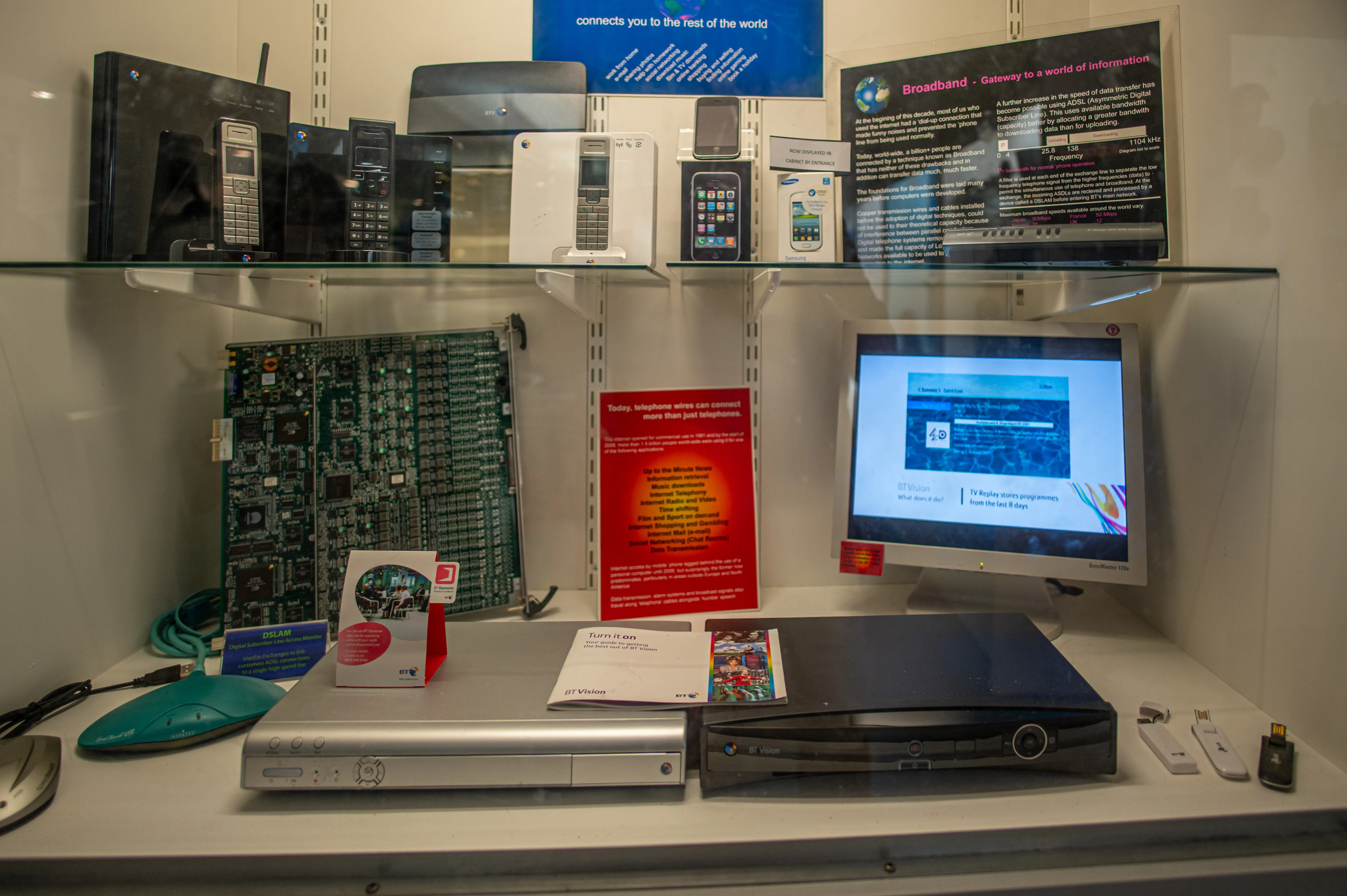 A museum display case with various telecommunications equipment, including vintage mobile phones, a computer circuit board, broadband modems, and a computer monitor displaying information about broadband.