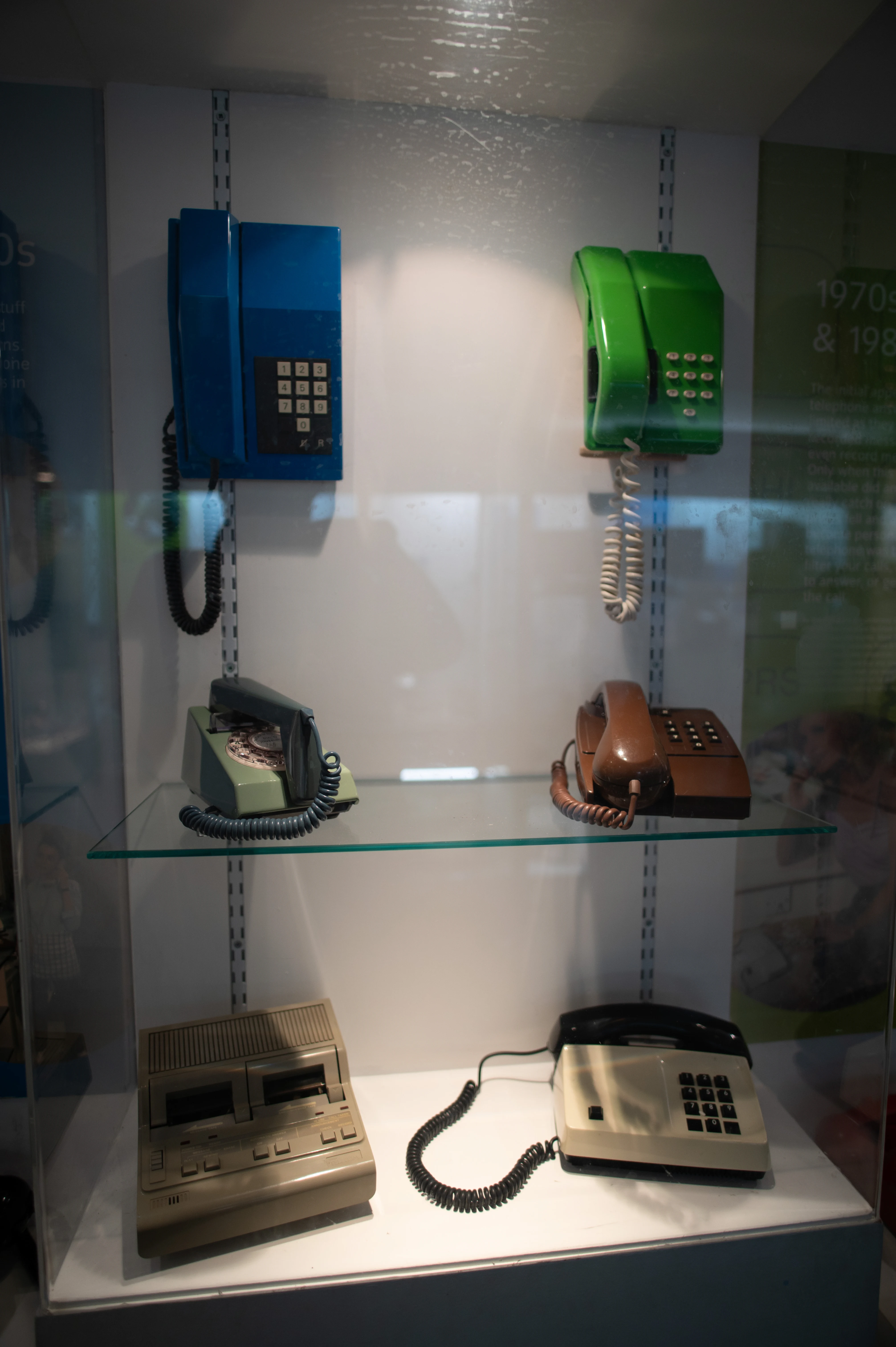 A display case containing a collection of old telephones in different colours and styles, including rotary dial and push-button models, alongside a retro data storage device.