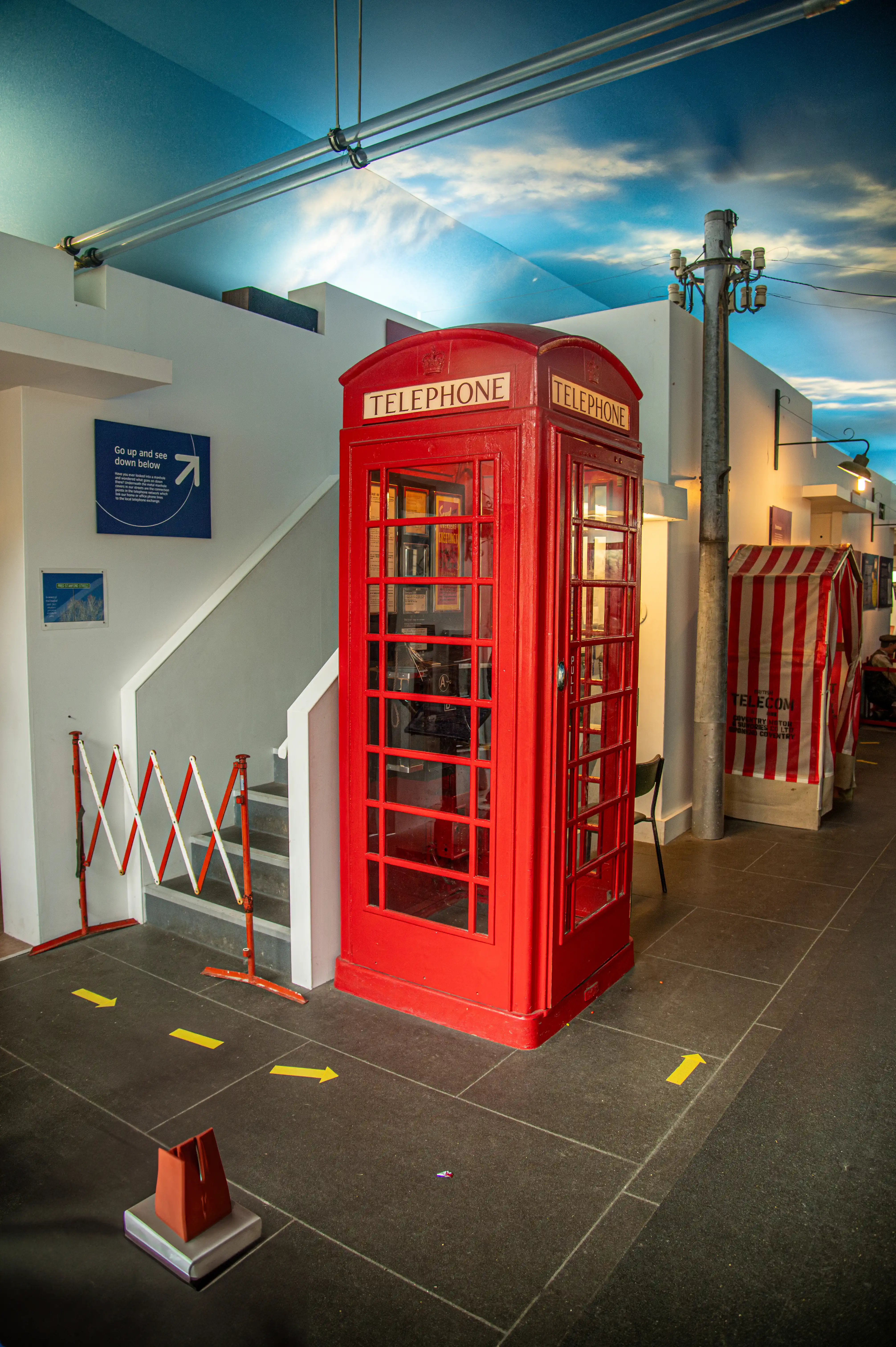 A bright red, classic British telephone box stands centrally in an exhibition hall. Above the telephone box, the ceiling is painted with a sky and clouds. Yellow arrows are marked on the dark floor, indicating a path. A staircase with a red and white barrier is visible to the left.