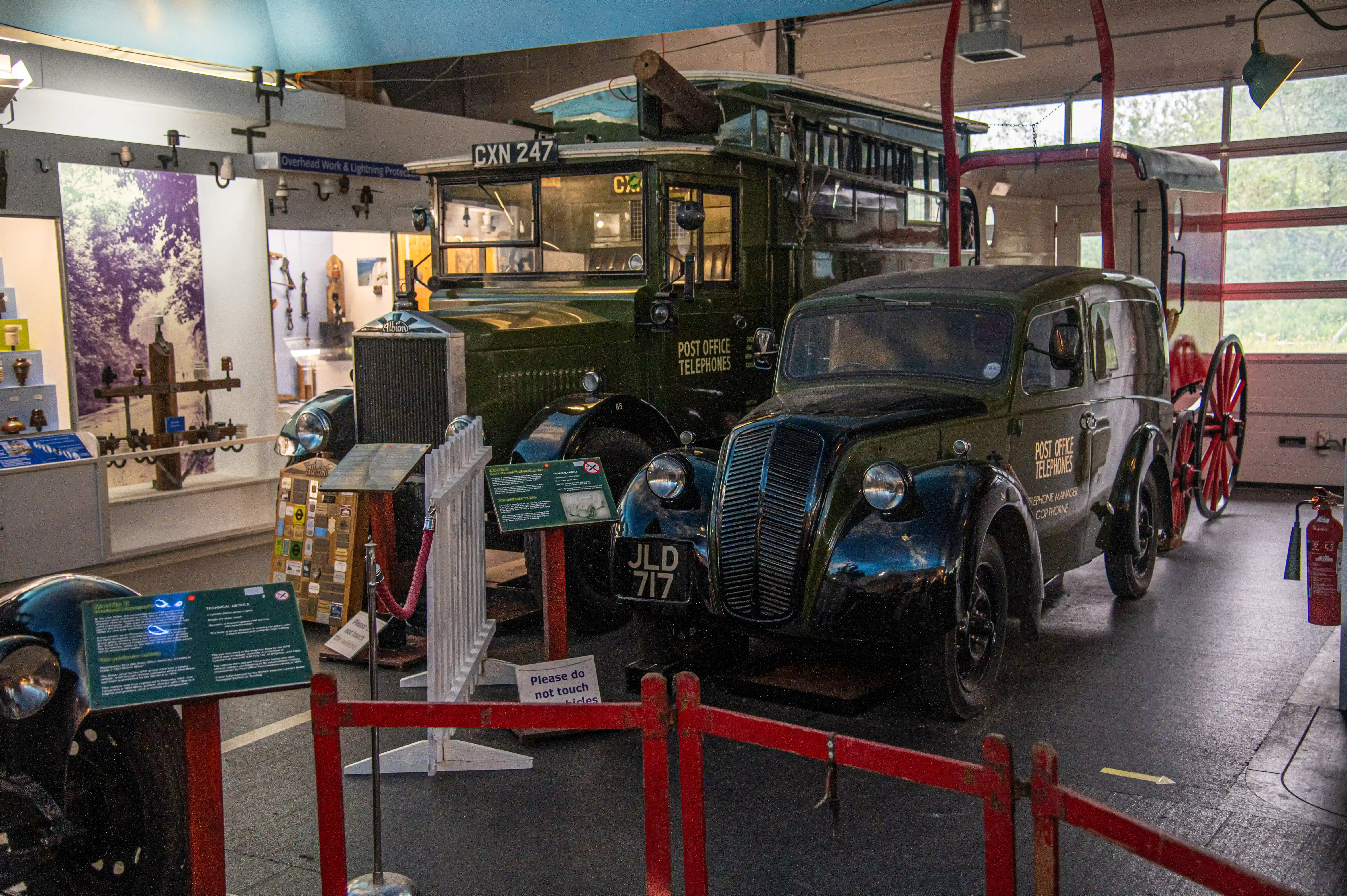 Two vintage dark green Post Office vehicles, a large bus and a smaller car, are parked side-by-side in a museum hall. The bus has 