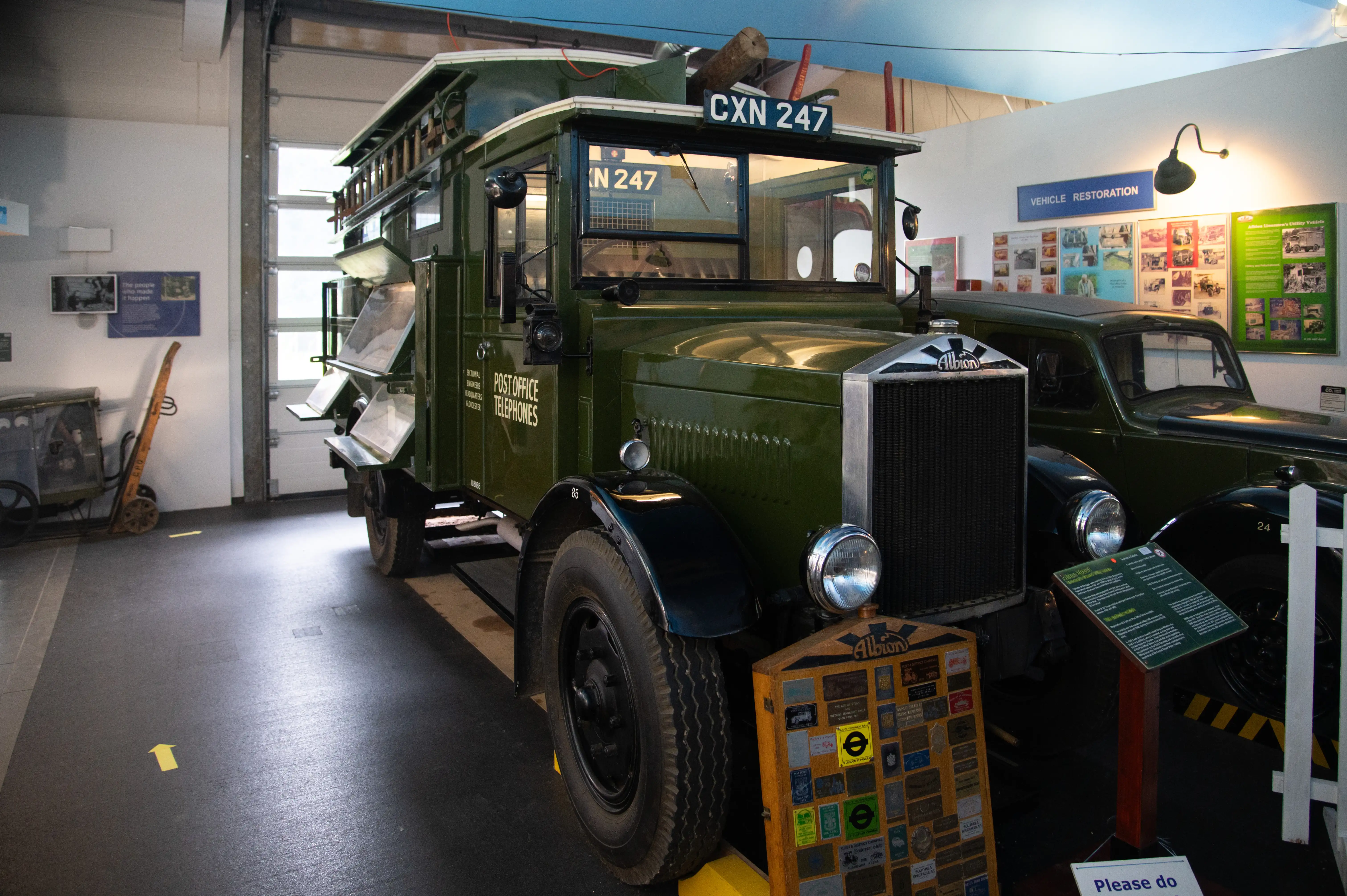 A vintage green Albion Post Office Telephones truck, with the registration CXN 247, is parked inside a museum or exhibition space. The truck has a ladder on its roof and is surrounded by other exhibits, including information boards and a display of badges.