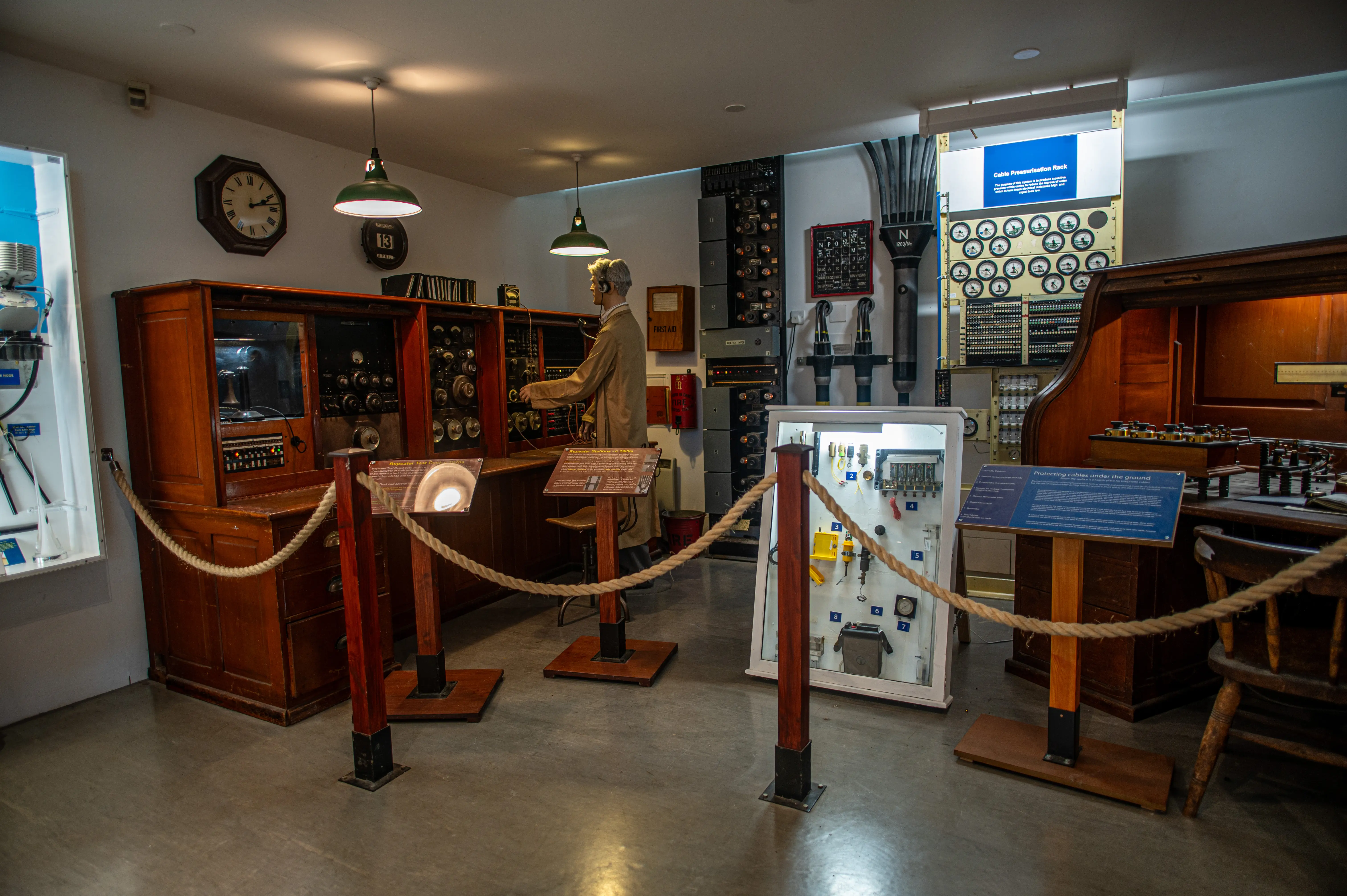 A dimly lit museum display of a vintage telecommunications room, with a mannequin dressed as an operator standing at a large wooden control desk covered in dials, switches, and monitoring equipment. In the background, there are racks of electronic components, clocks on the wall, and informational displays.