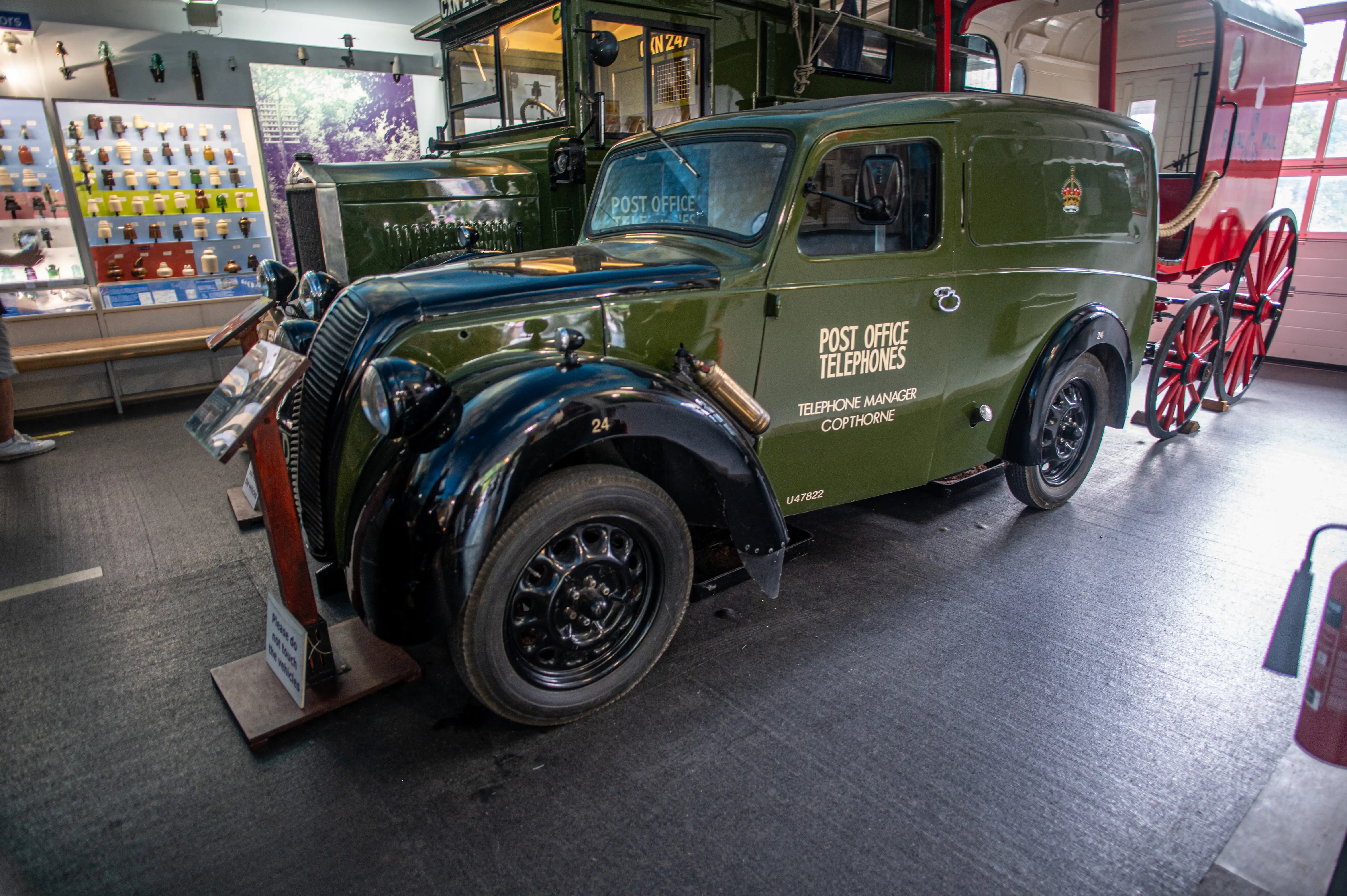 A vintage green Post Office van with black fenders and wheels, parked indoors. The side of the van displays &laquo;POST OFFICE TELEPHONES&raquo