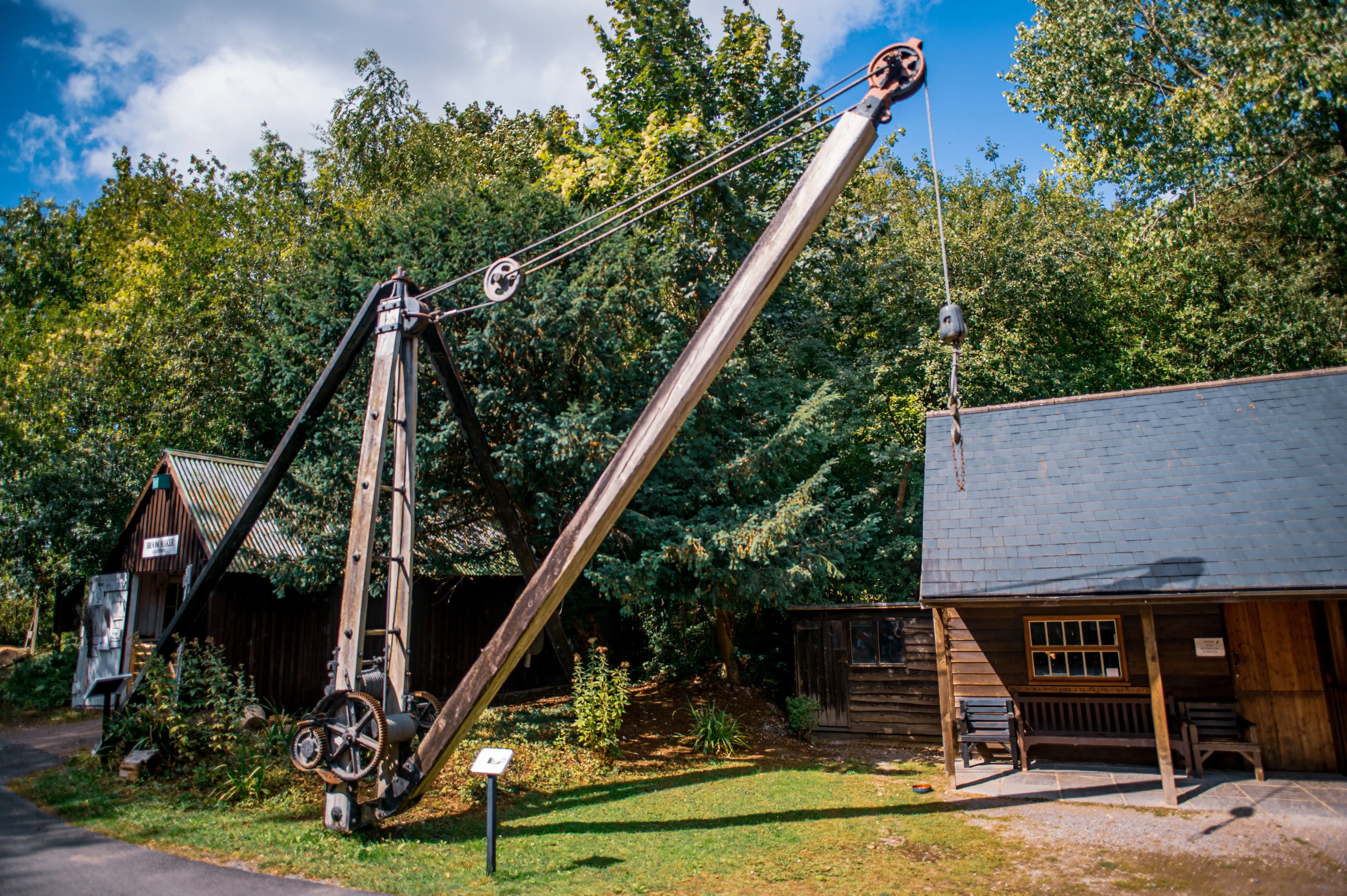 A vintage wooden crane with a long boom is positioned outdoors in front of two small wooden buildings amidst dense foliage and under a partly cloudy sky.