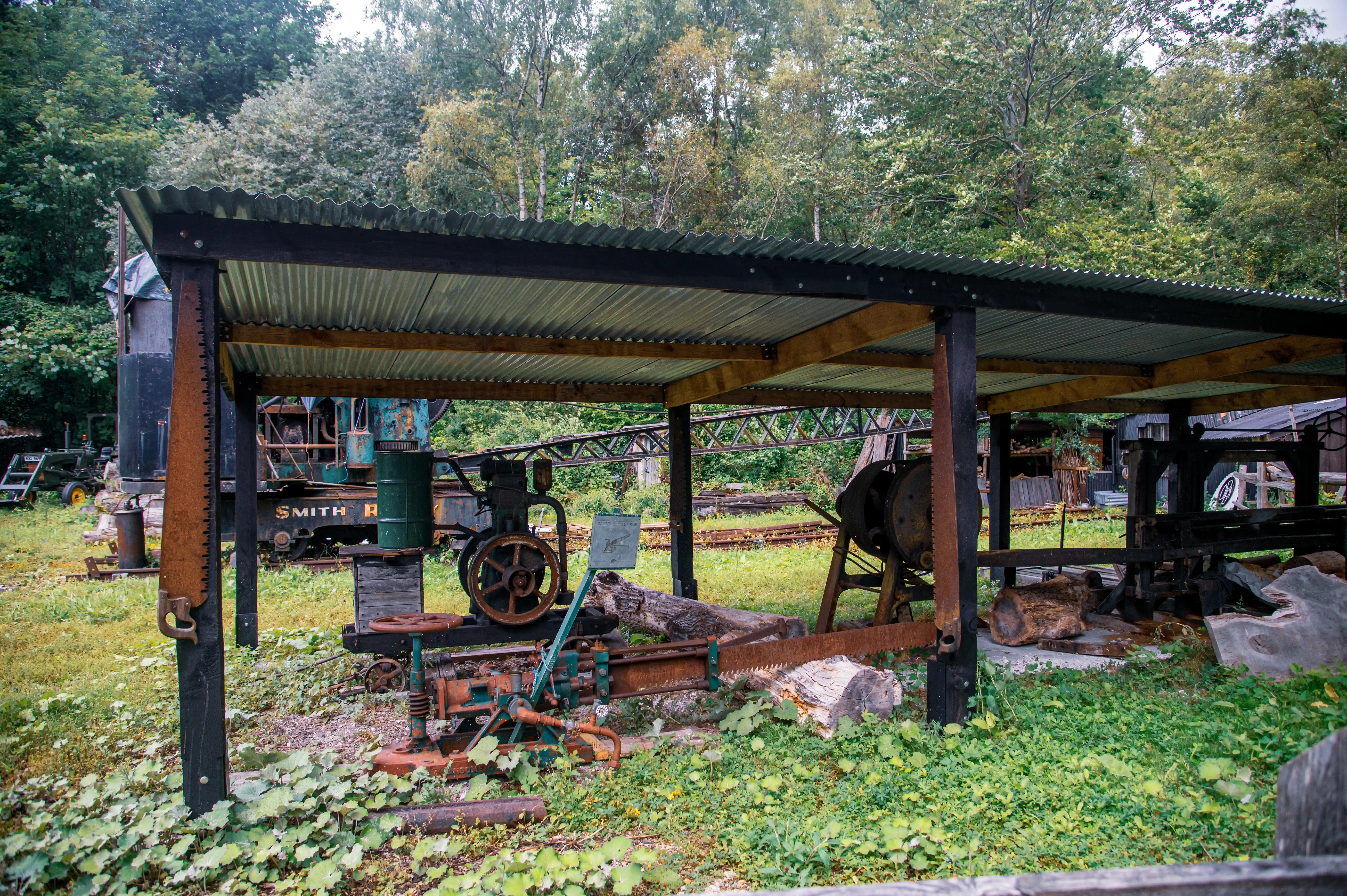 An outdoor view of a rustic, weathered sawmill with various old machines, large saws, and cut logs situated under a simple open-sided shelter. Trees and green foliage surround the area, creating a sense of a bygone era.