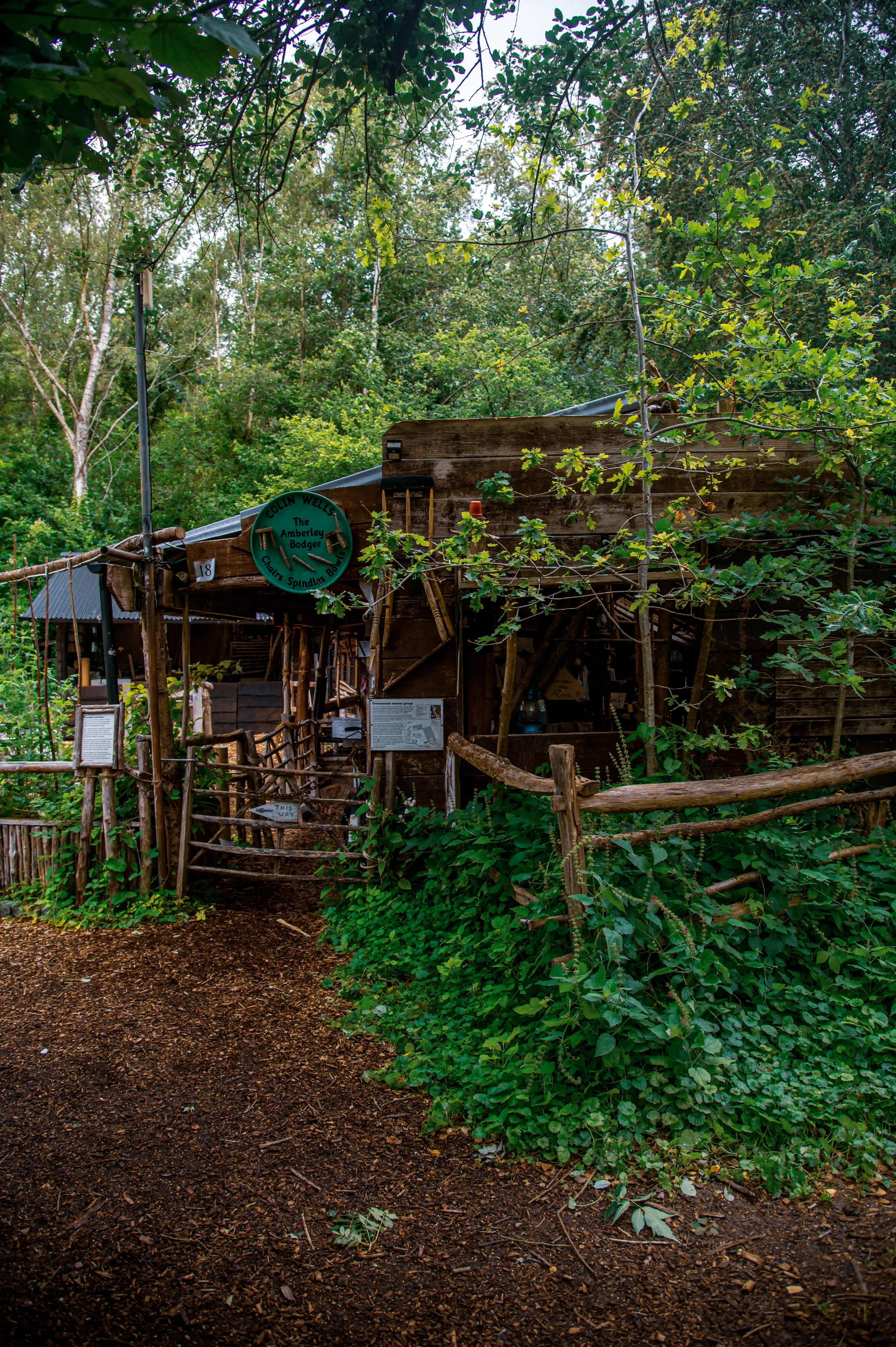 A rustic wooden workshop building situated within a dense, green forest setting, with a wooden gate and fence in the foreground and a circular sign above the entrance.