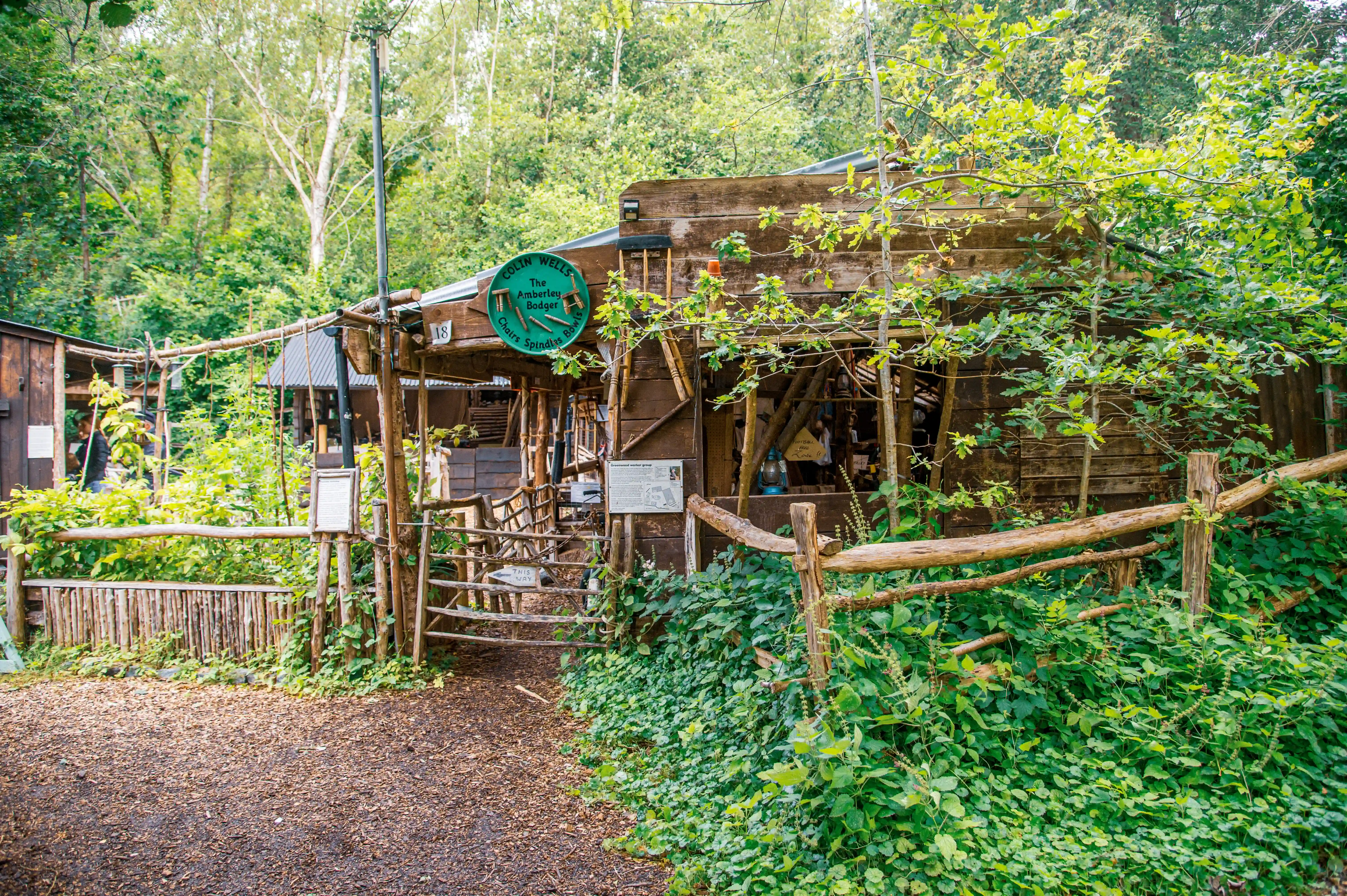 A rustic wooden workshop surrounded by trees and greenery, with a wooden fence and gate.