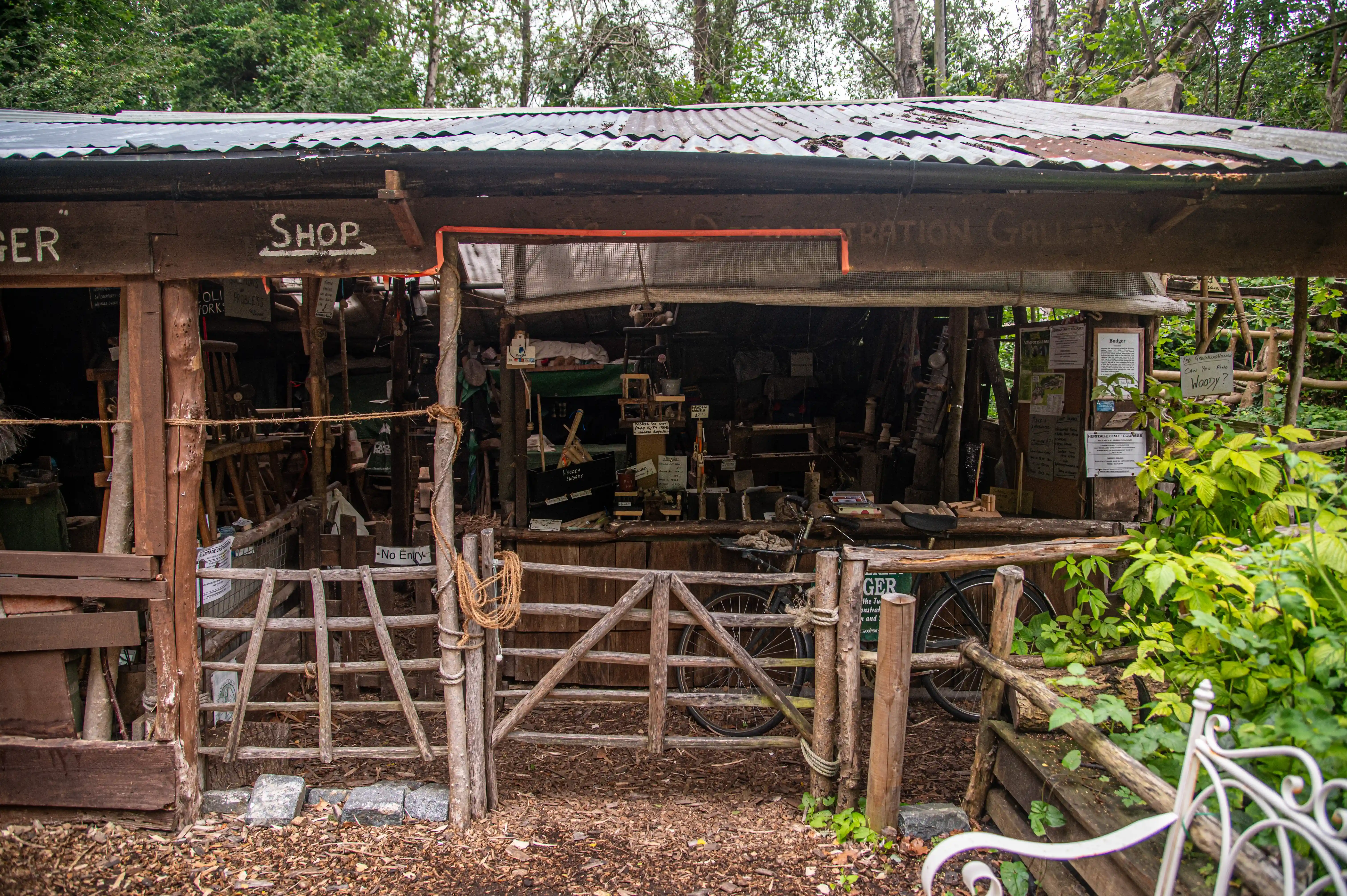 A rustic wooden workshop surrounded by trees and greenery, with a wooden fence and gate.