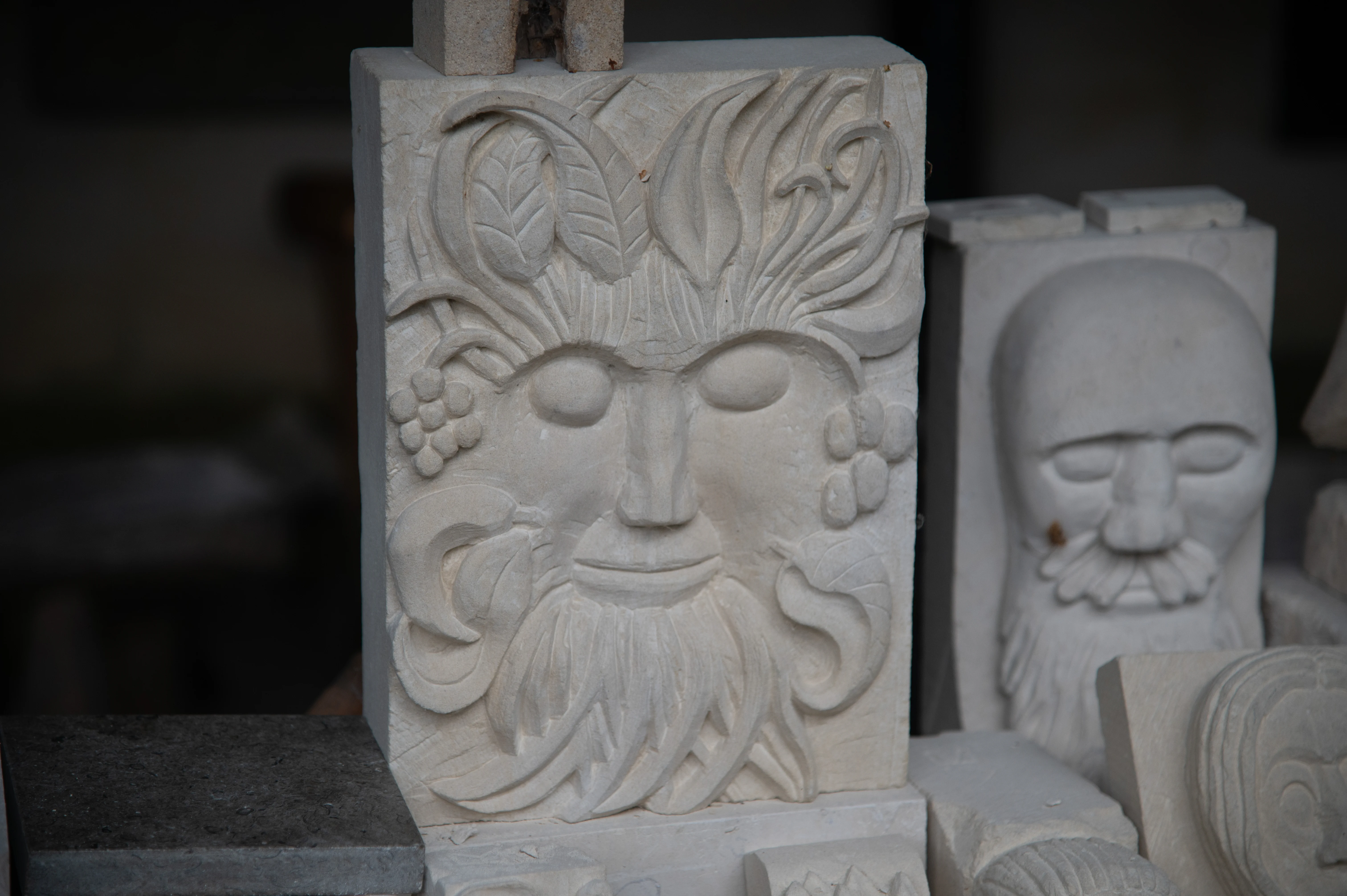 A stone relief carving of a face with a long beard, leaves, and grapes. Another stone carving of a face is blurred in the background.