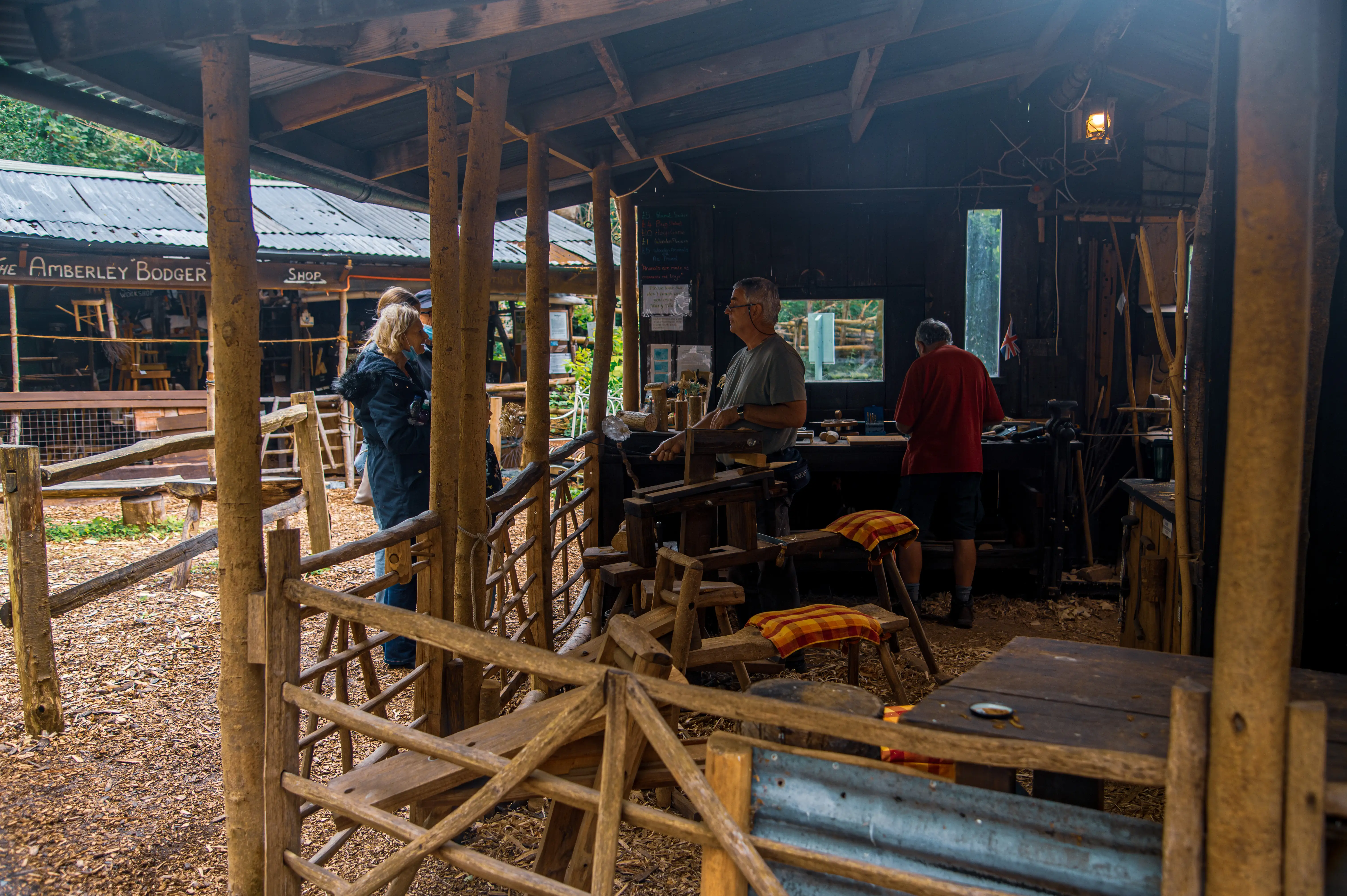 A woodworking demonstration inside a rustic wooden workshop, with three people visible and tools laid out on workbenches.