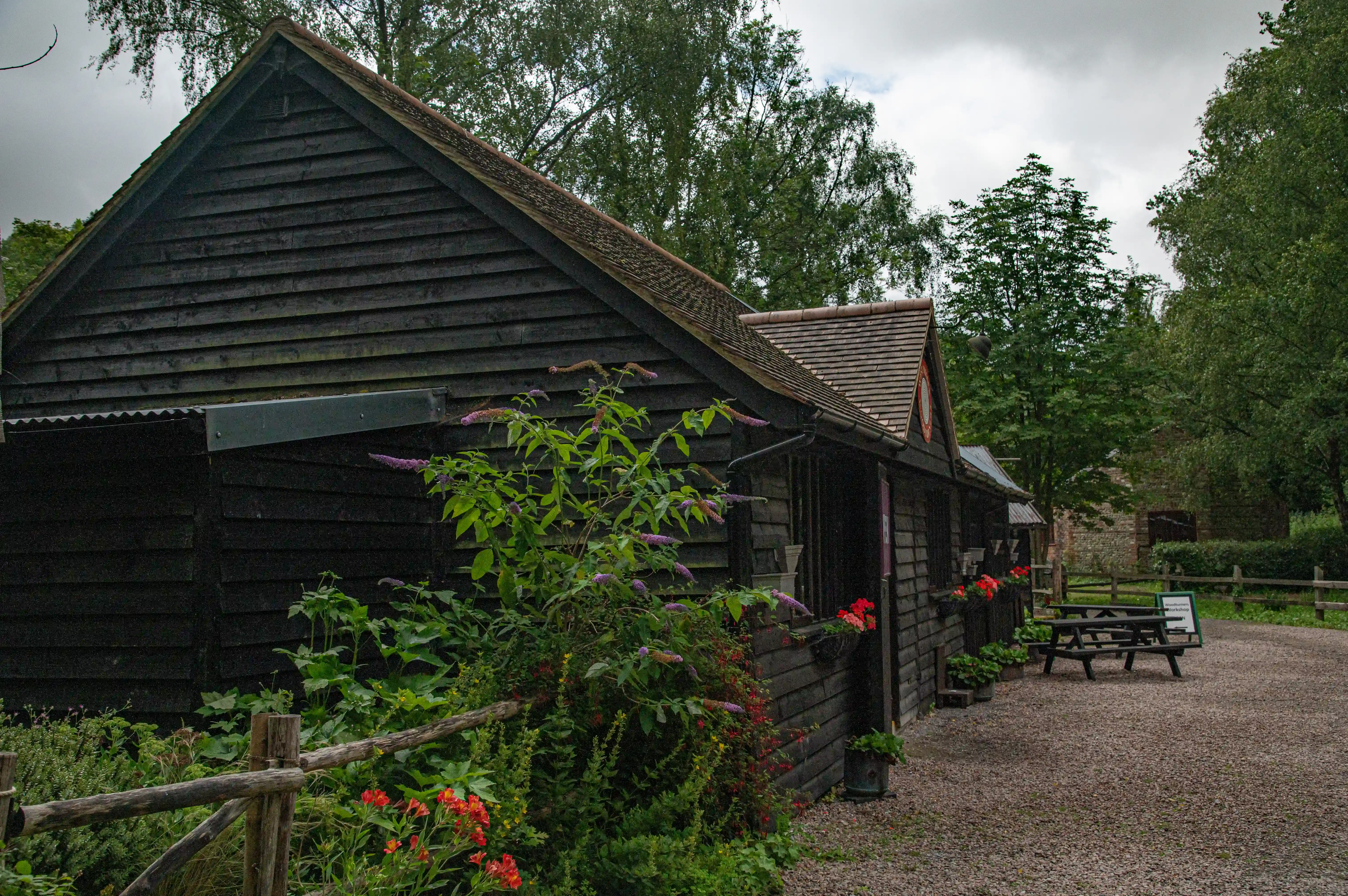 A row of dark wooden buildings with pitched roofs and flower boxes filled with red and purple blooms. A gravel path runs in front of the buildings, with a wooden fence and trees in the background