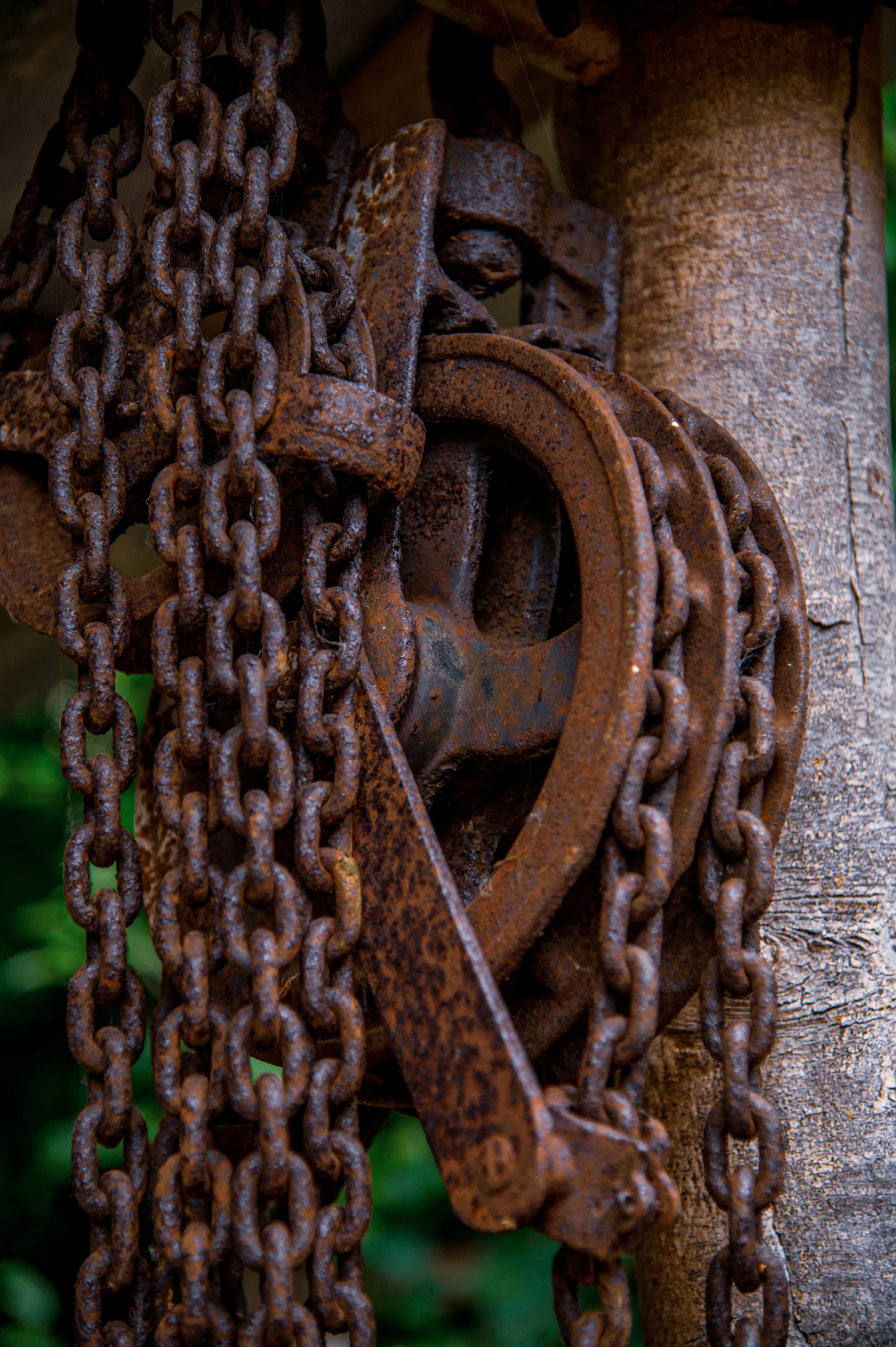 Close-up of a rusty metal pulley wheel wrapped with heavy chains, mounted next to a textured wooden post.