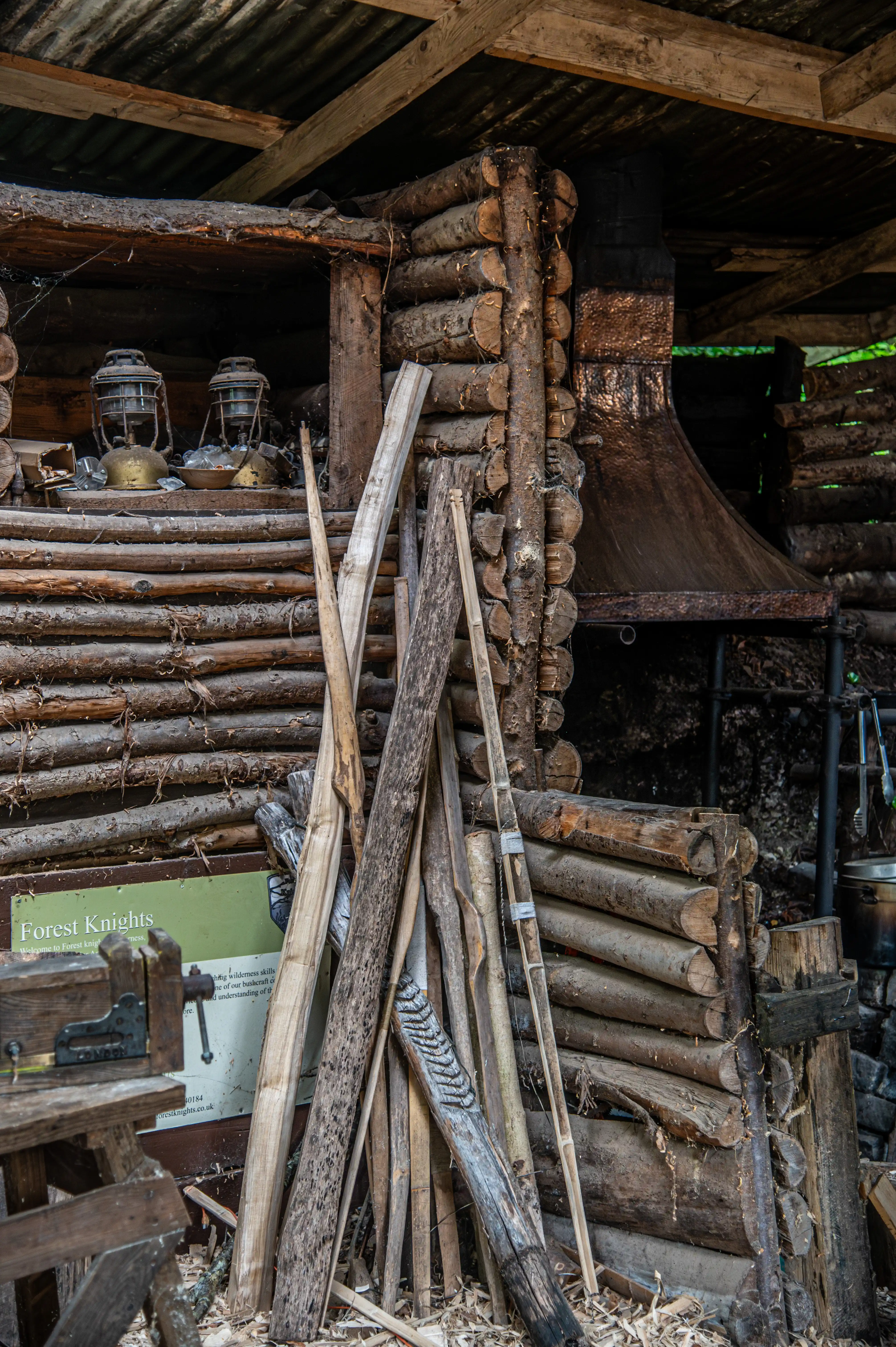 A close-up shot shows the rough-hewn interior and exterior of a log cabin-style structure. Two vintage oil lanterns are visible inside. Various wooden poles and branches are stacked against the logs, and a sign with the words 