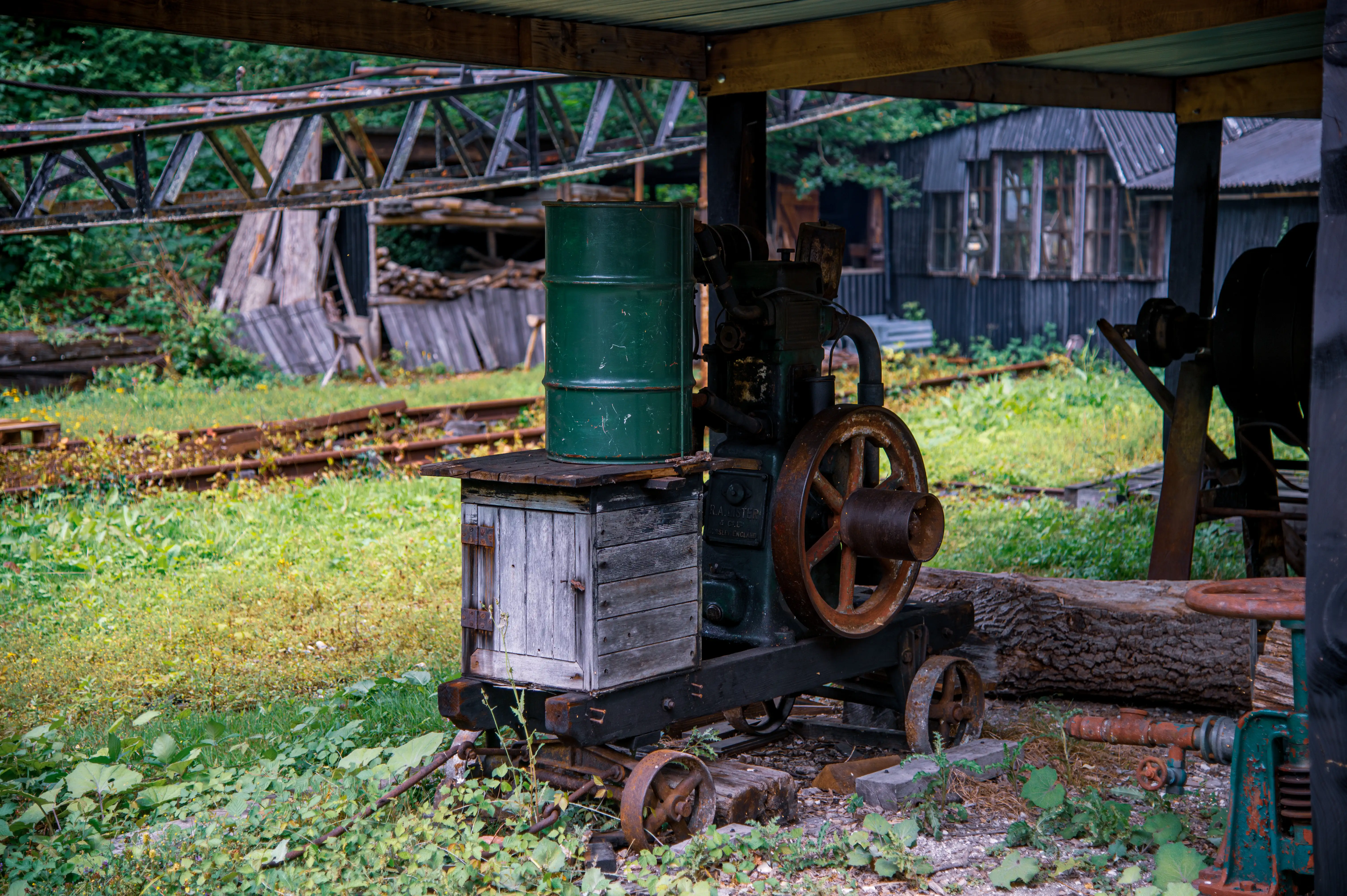 A close-up view of a vintage industrial engine with a large rusty flywheel, a green barrel, and a wooden box, all under a wooden shelter. Overgrown railway tracks and derelict buildings are visible in the background.