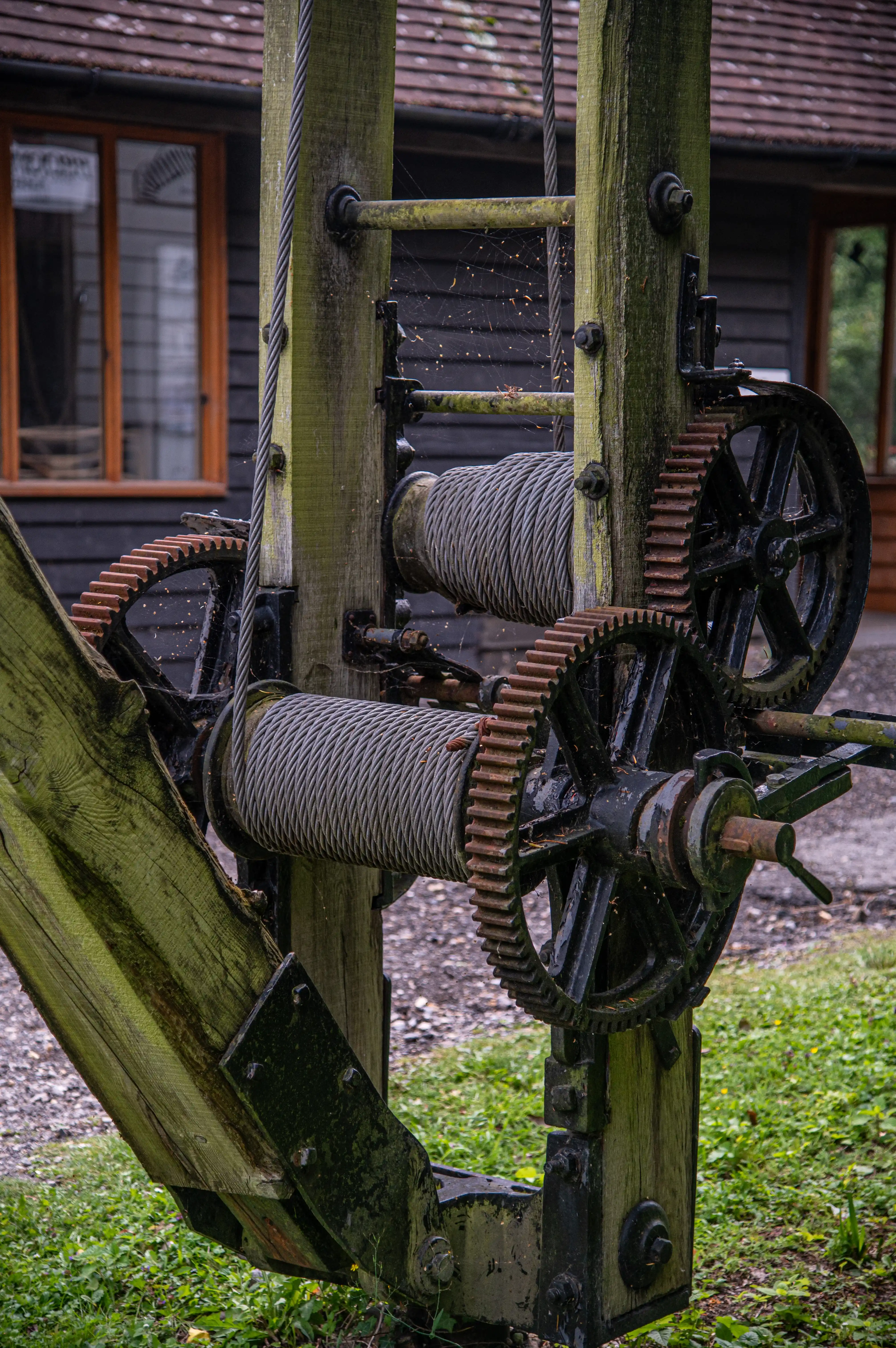 Detailed shot of an old metal and wood winch with two large wound cables and a pair of cogged gears, one partially rusted, set against a rustic building.