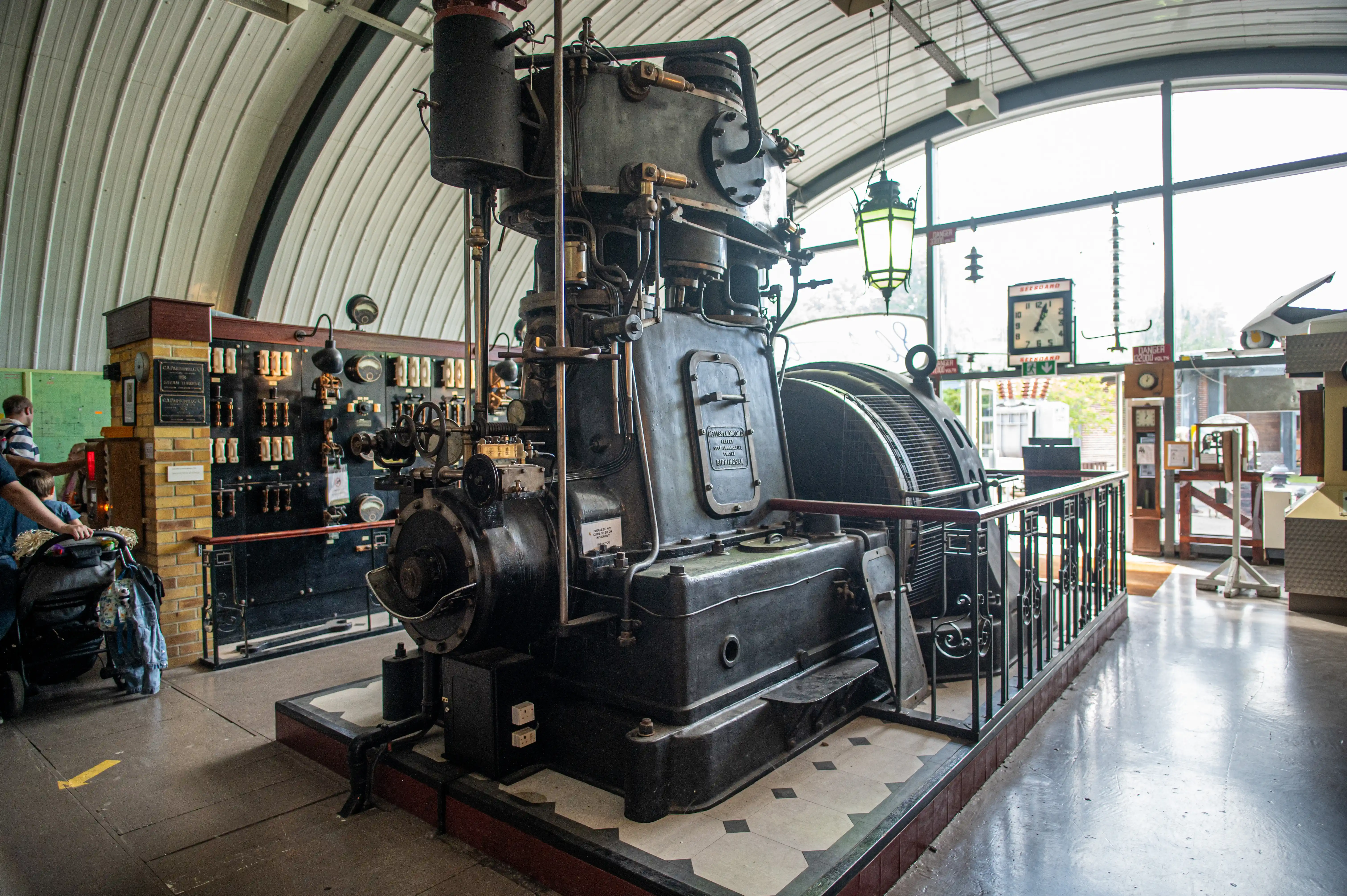 A large, dark, industrial steam turbine with intricate pipework and gauges is on display in a museum. To its left is a vintage electrical control panel with numerous switches and meters. A black metal railing surrounds the turbine. The hall has a high, curved, corrugated metal ceiling and large windows, with a green lantern hanging from above.