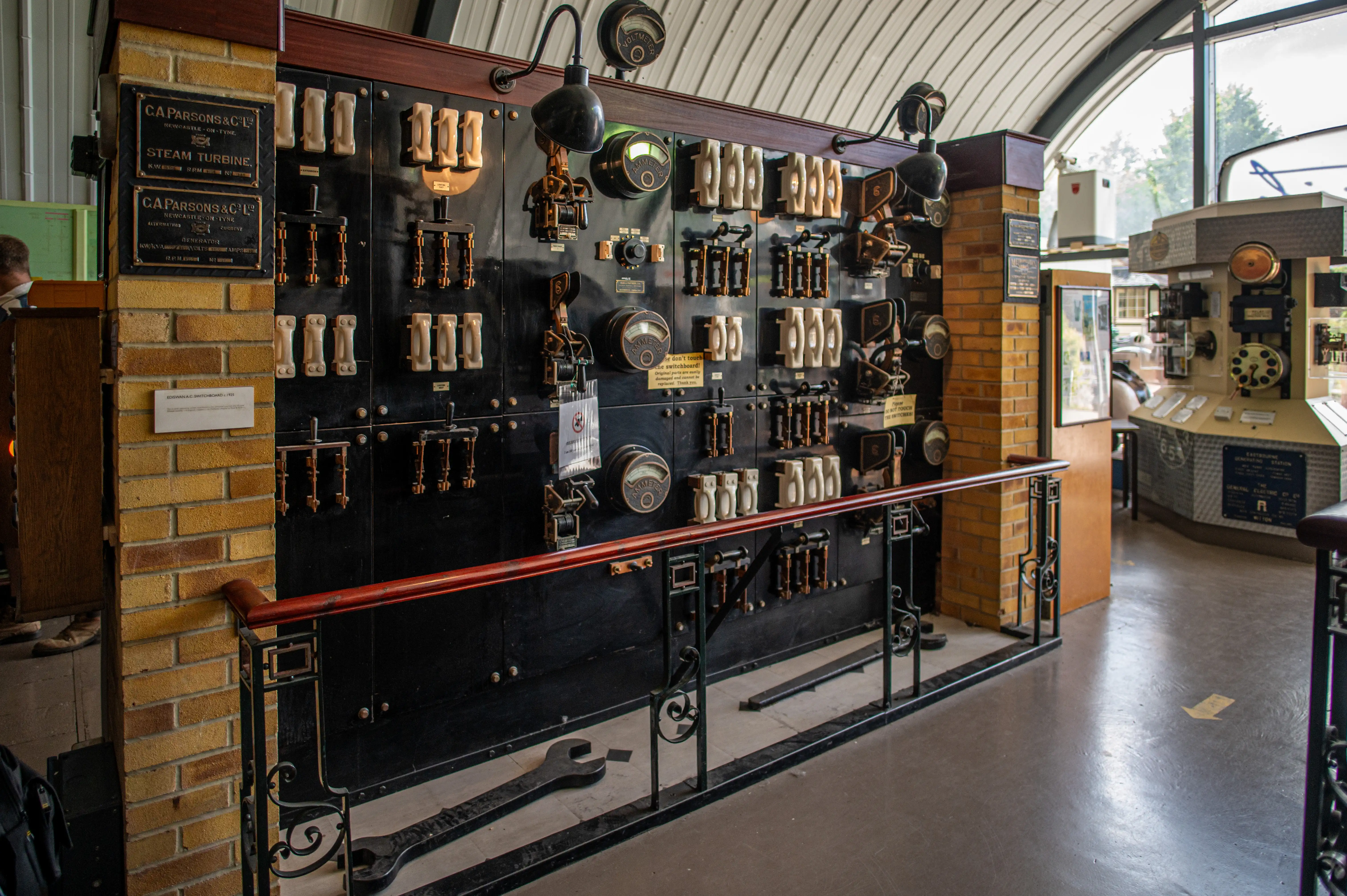 A large, dark, vintage electrical switchboard with rows of white ceramic switches and brass levers, mounted on a black panel. Several analogue meters, including voltmeters and ammeters, are integrated into the panel. A brick pillar with C.A. Parsons & Co. plaques is to the left, and a red wooden handrail runs across the front of the exhibit.