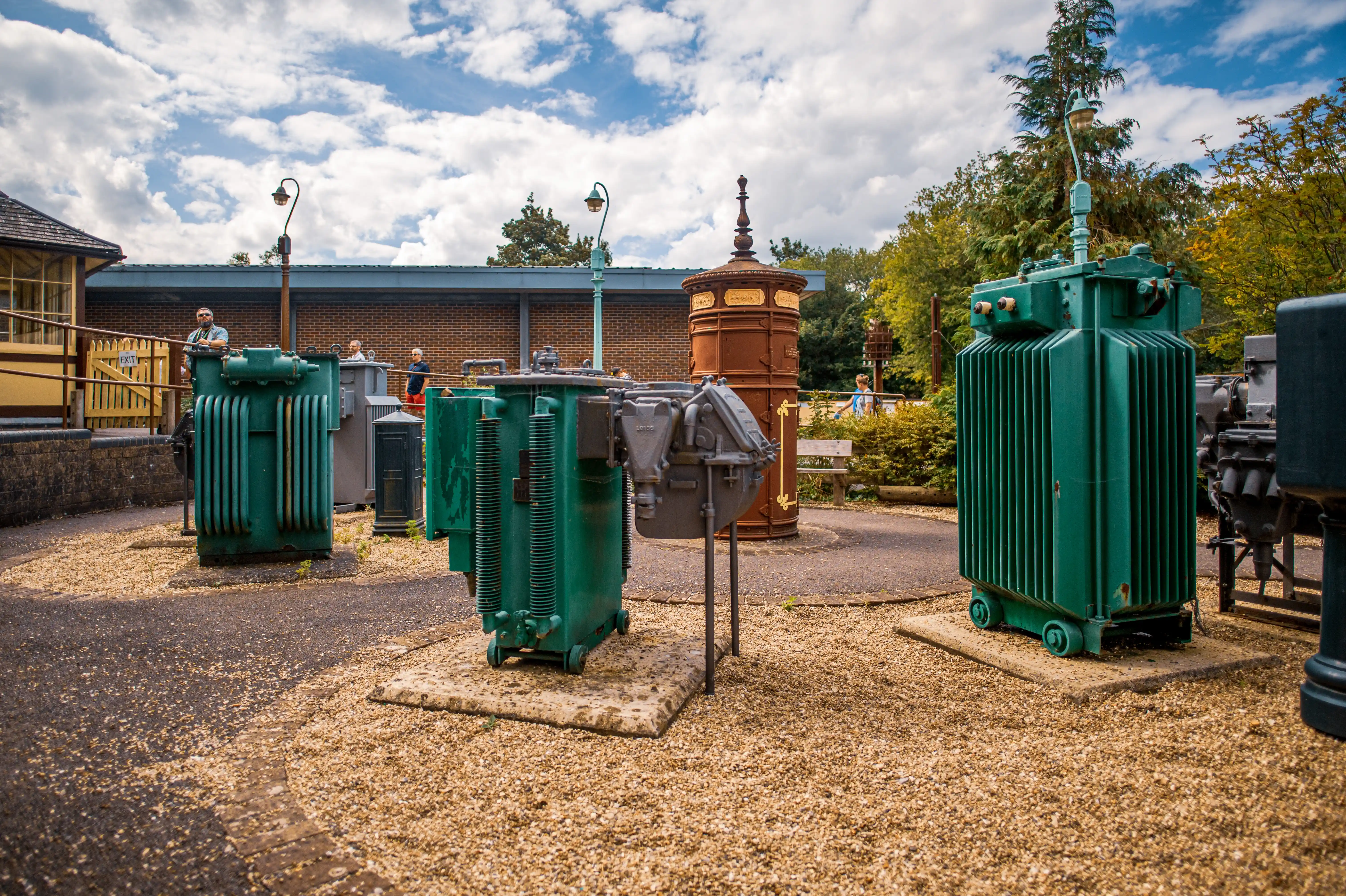 Several large, vintage green electrical transformers are positioned outdoors on gravel. A brick building with a yellow section and an 'EXIT' sign is in the background, under a blue sky with white clouds.