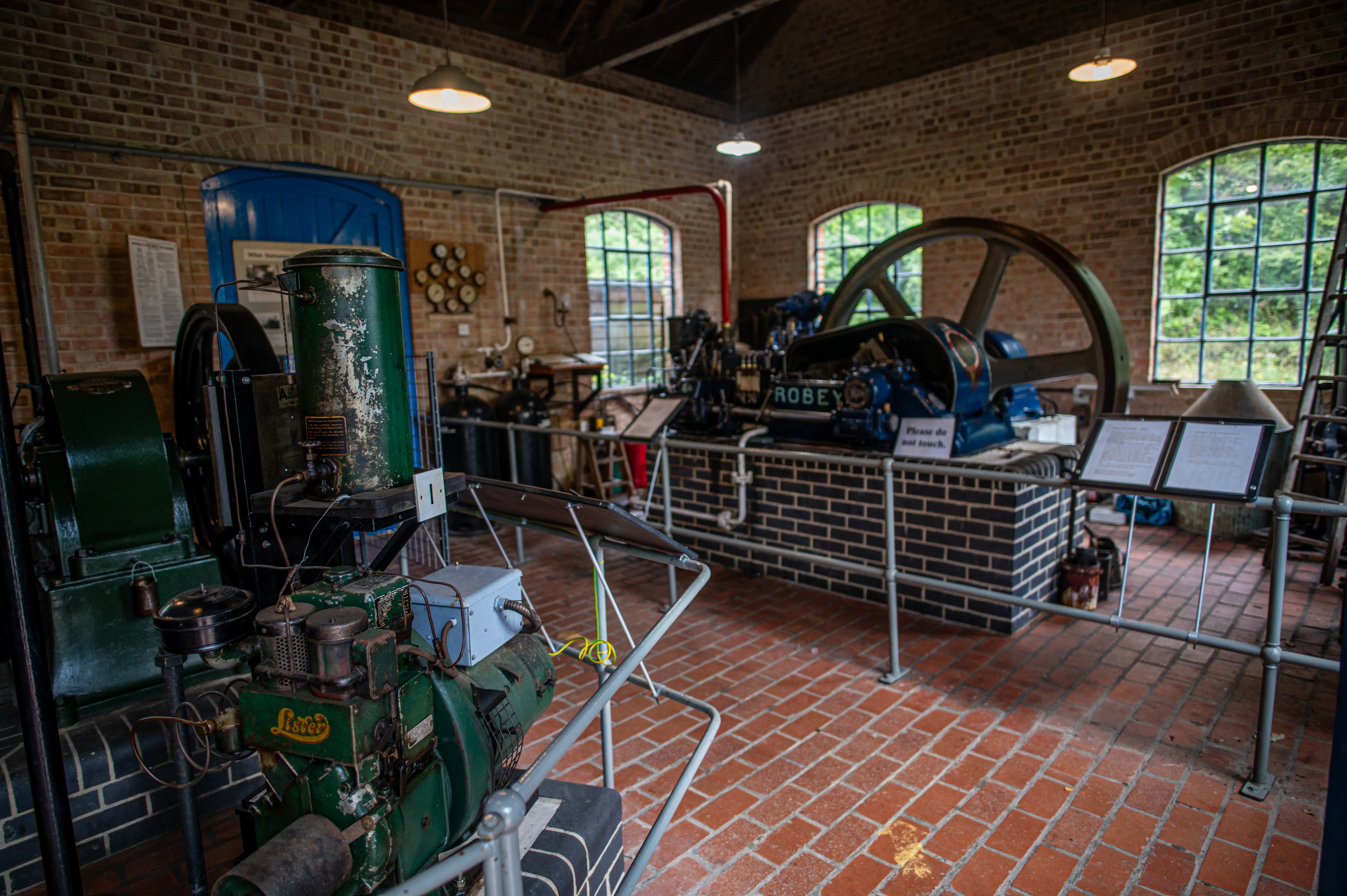Inside a museum, several vintage industrial engines are on display. In the foreground, a green Lister engine is visible, with a larger blue Robey engine and its large flywheel in the background. Brick walls, arched windows with greenery outside, and a red-tiled floor are present.