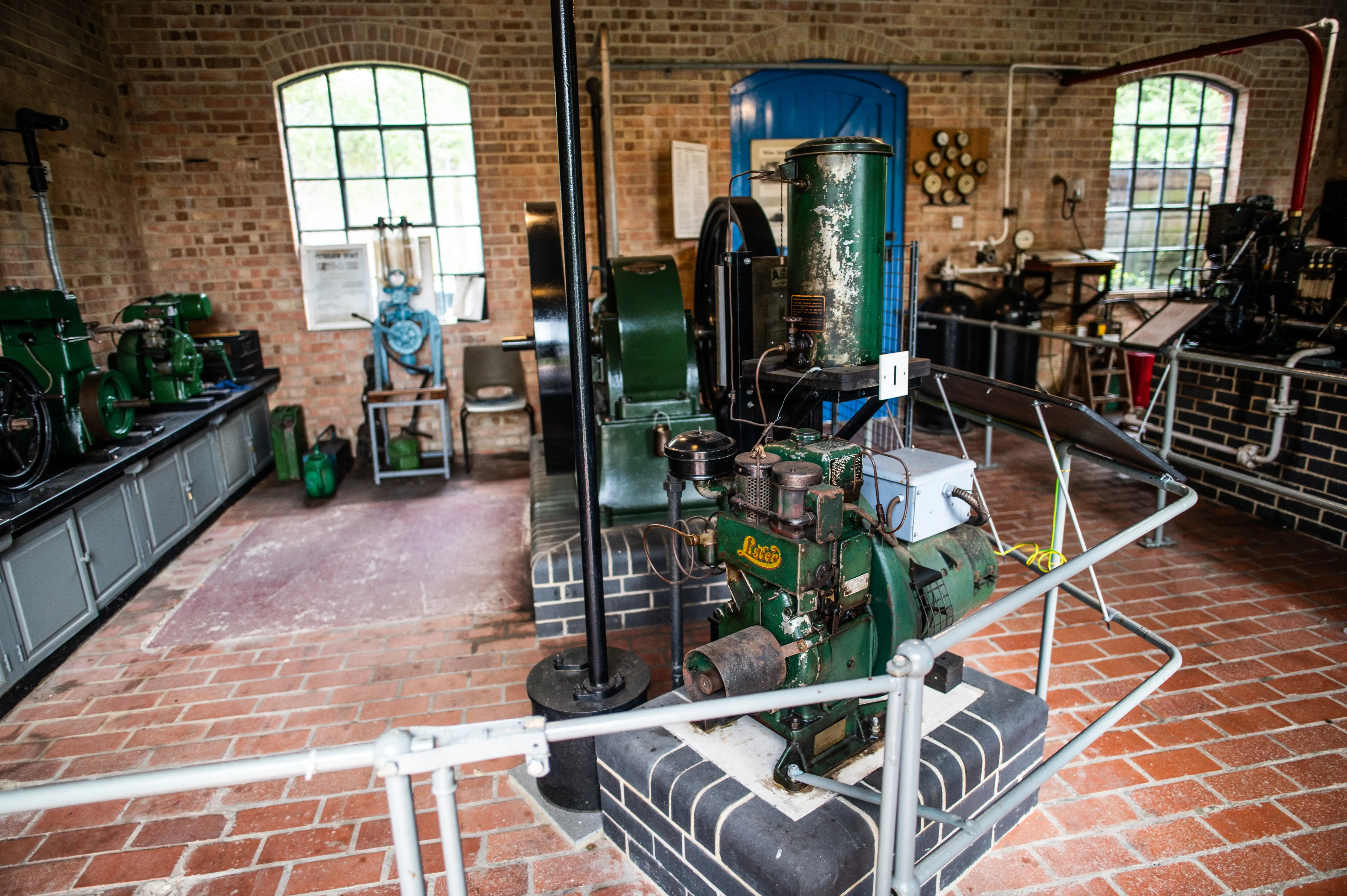 Inside a museum, several vintage industrial engines are on display. In the foreground, a green Lister engine is visible, with a larger blue Robey engine and its large flywheel in the background. Brick walls, arched windows with greenery outside, and a red-tiled floor are present.