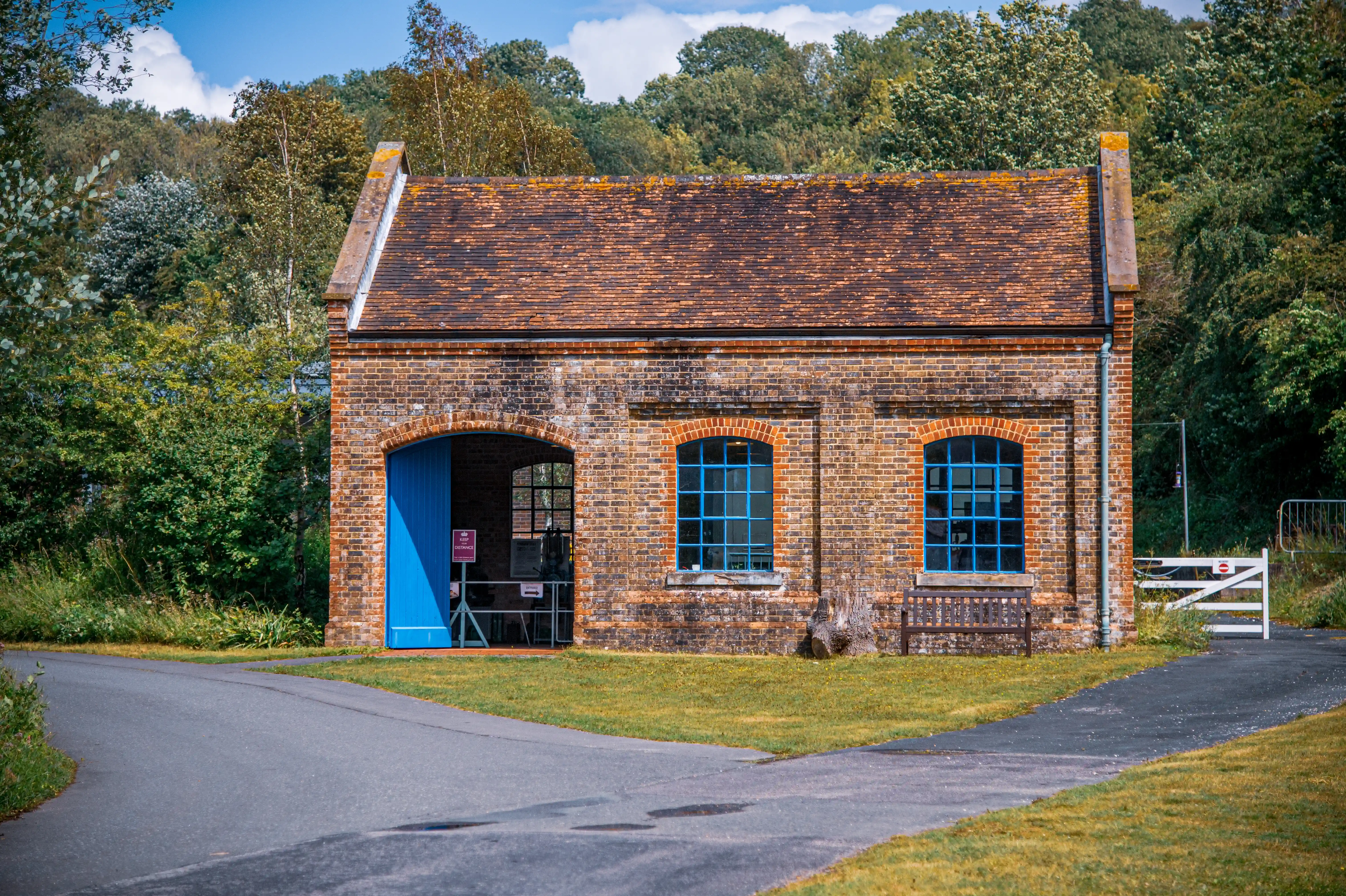 Small brick building with tiled roof, blue arched doors and windows, wooden bench, and surrounding greenery.