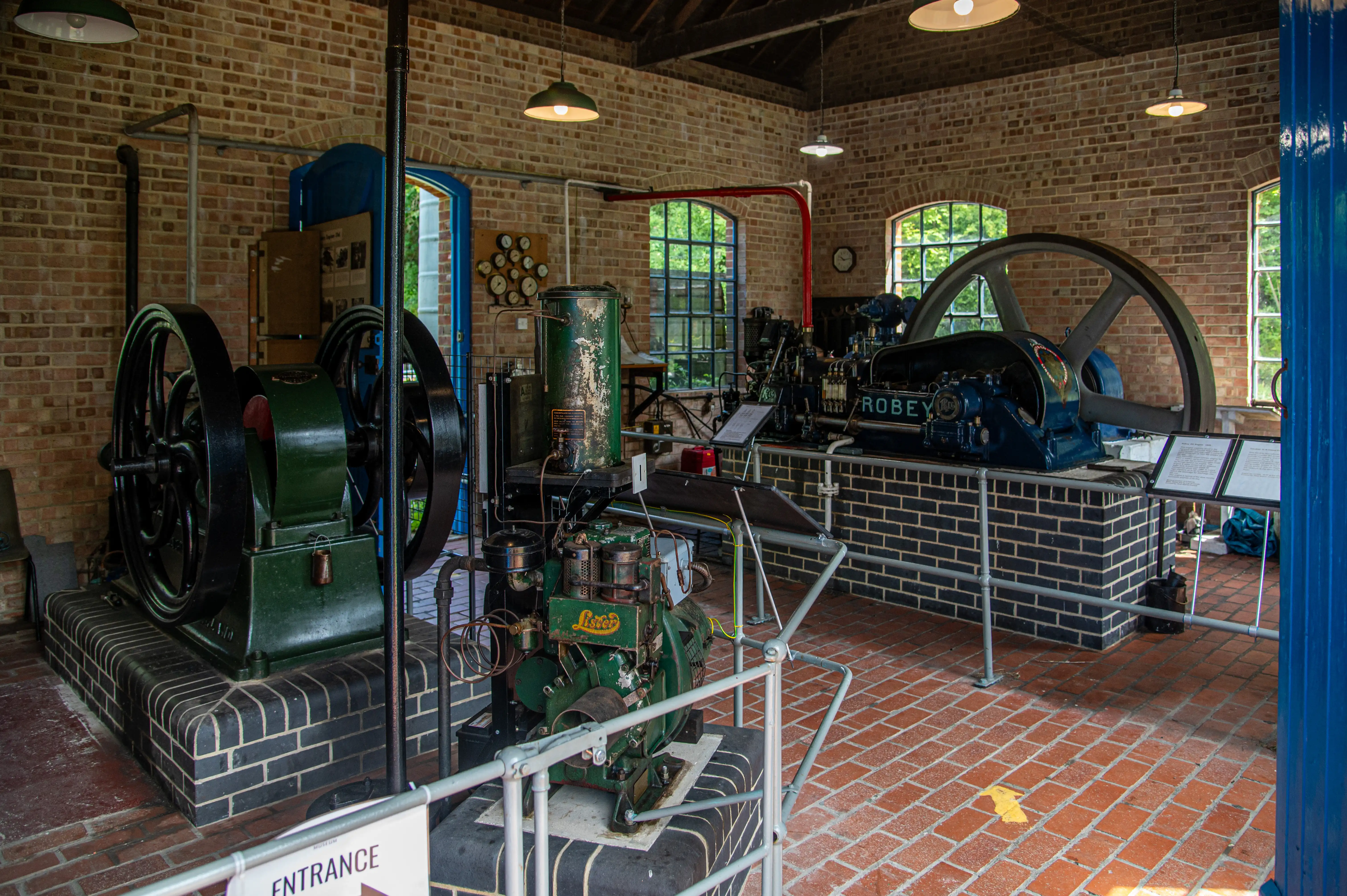 Interior view of a museum exhibit featuring several large, vintage industrial engines. In the foreground to the left is a green engine with large black flywheels. To the right, a blue engine with the name 