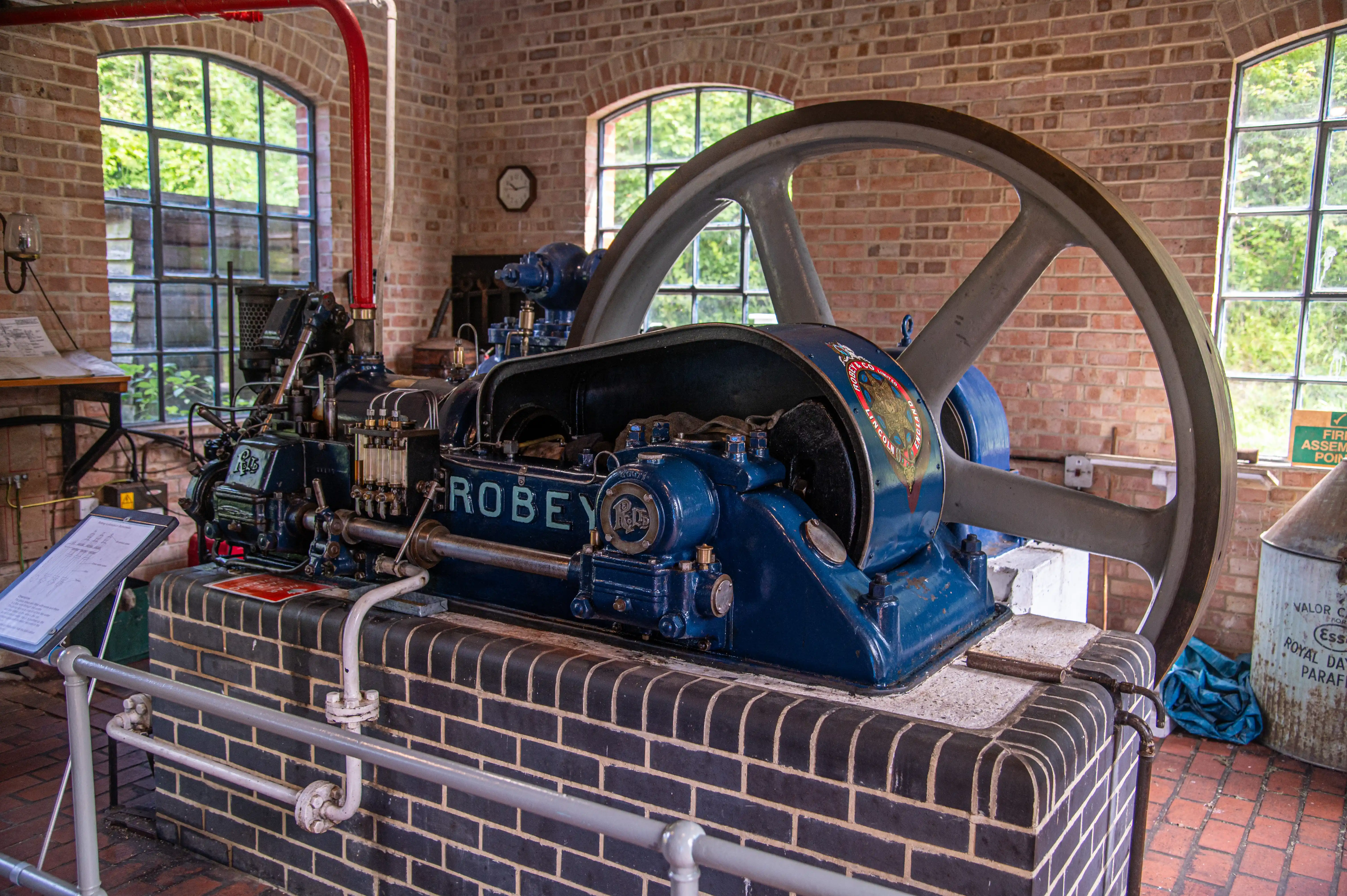 A detailed photograph of a blue Robey steam engine with a large grey flywheel, situated on a brick plinth in a room with brick walls and large windows looking out to greenery.