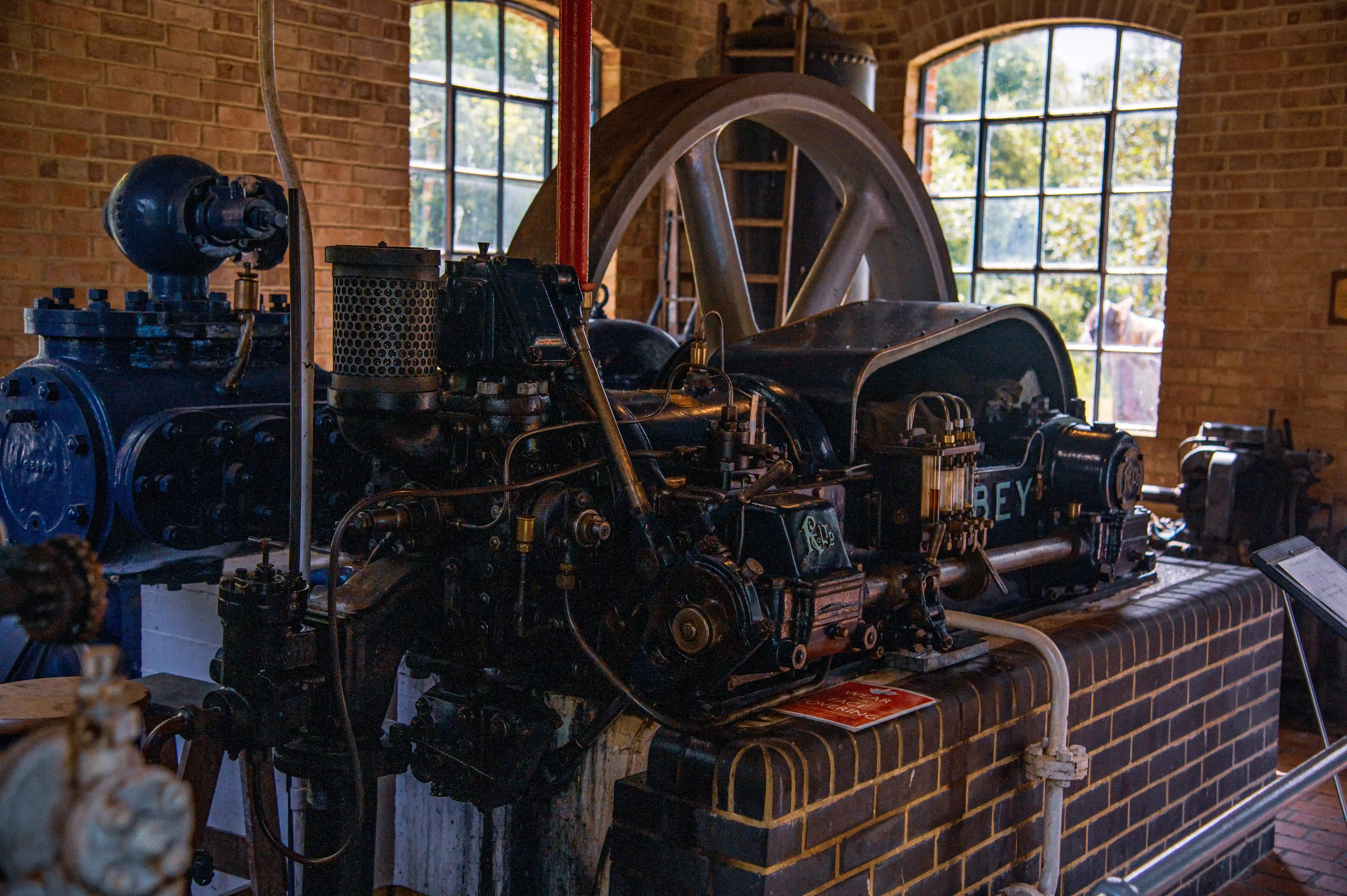 Close-up of a dark, complex vintage steam engine with a large flywheel visible in the background, set against a brick wall with large arched windows.