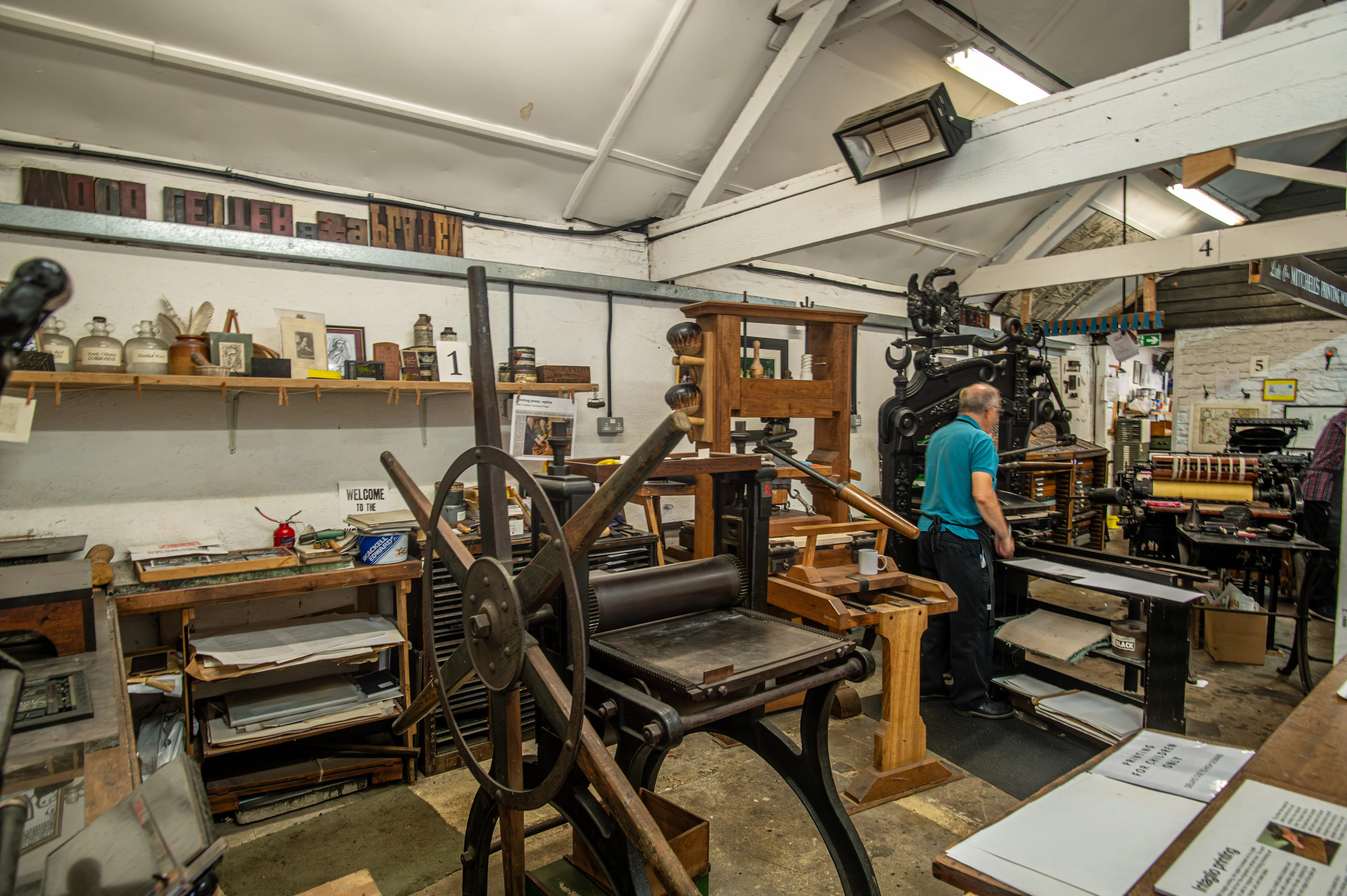Traditional printmaking workshop with historic presses, shelves of tools, and two people working with printing equipment.