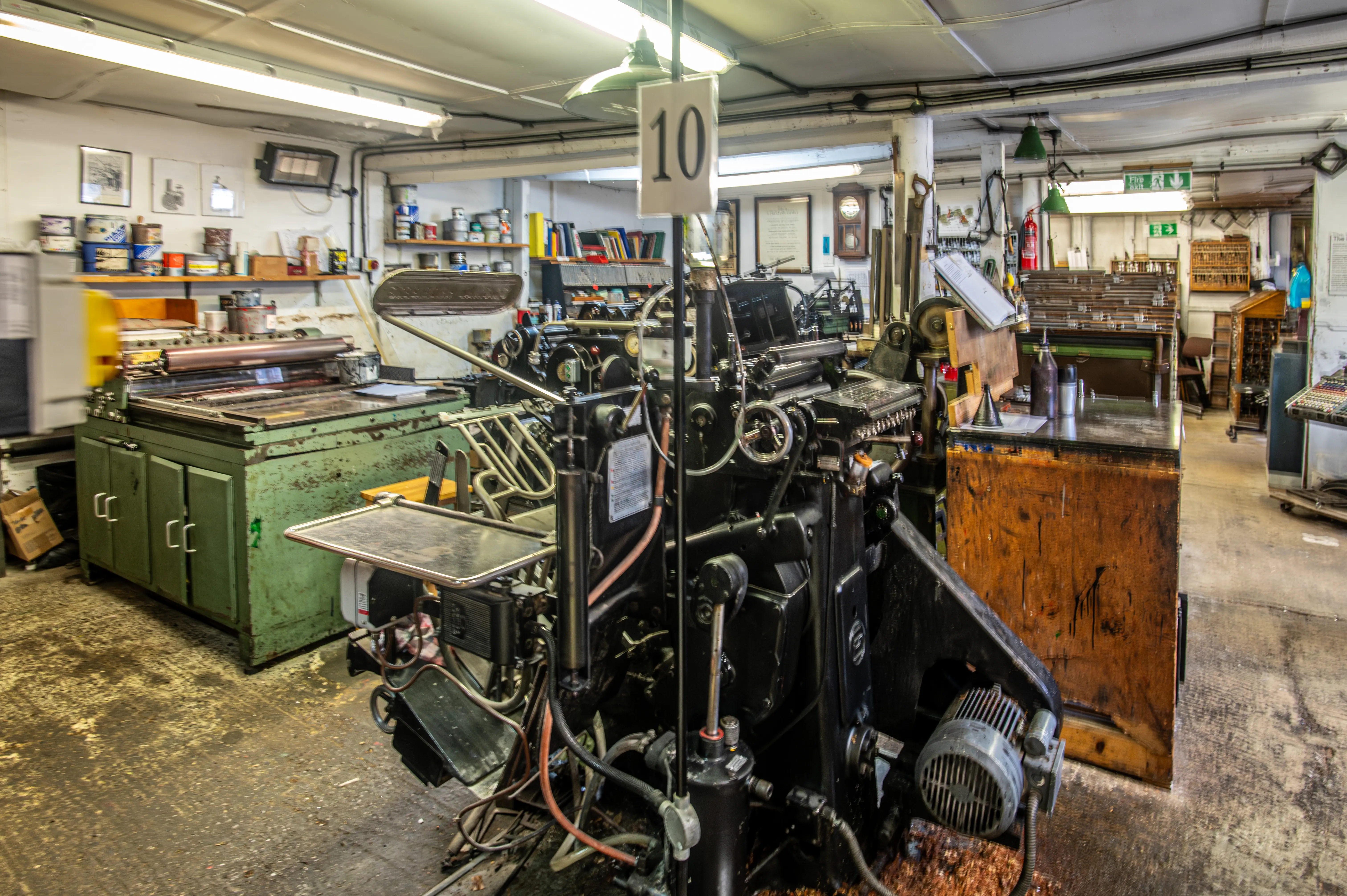 Large mechanical printing press in a traditional workshop with workbenches, cabinets, shelves of supplies, and additional printing equipment.