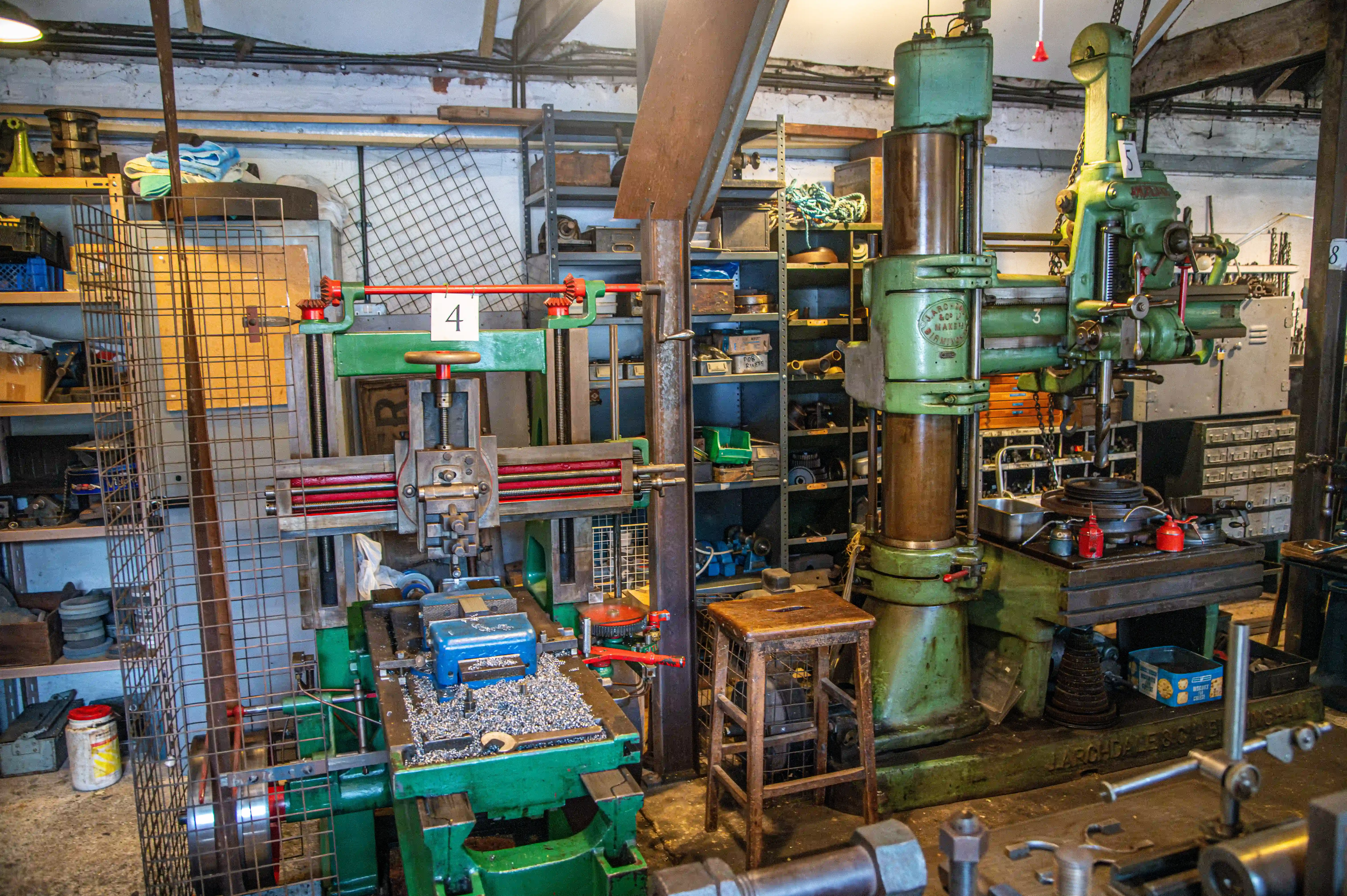 Green milling machine and radial drill press in a traditional workshop with shelves of tools and metal shavings on the workbench.