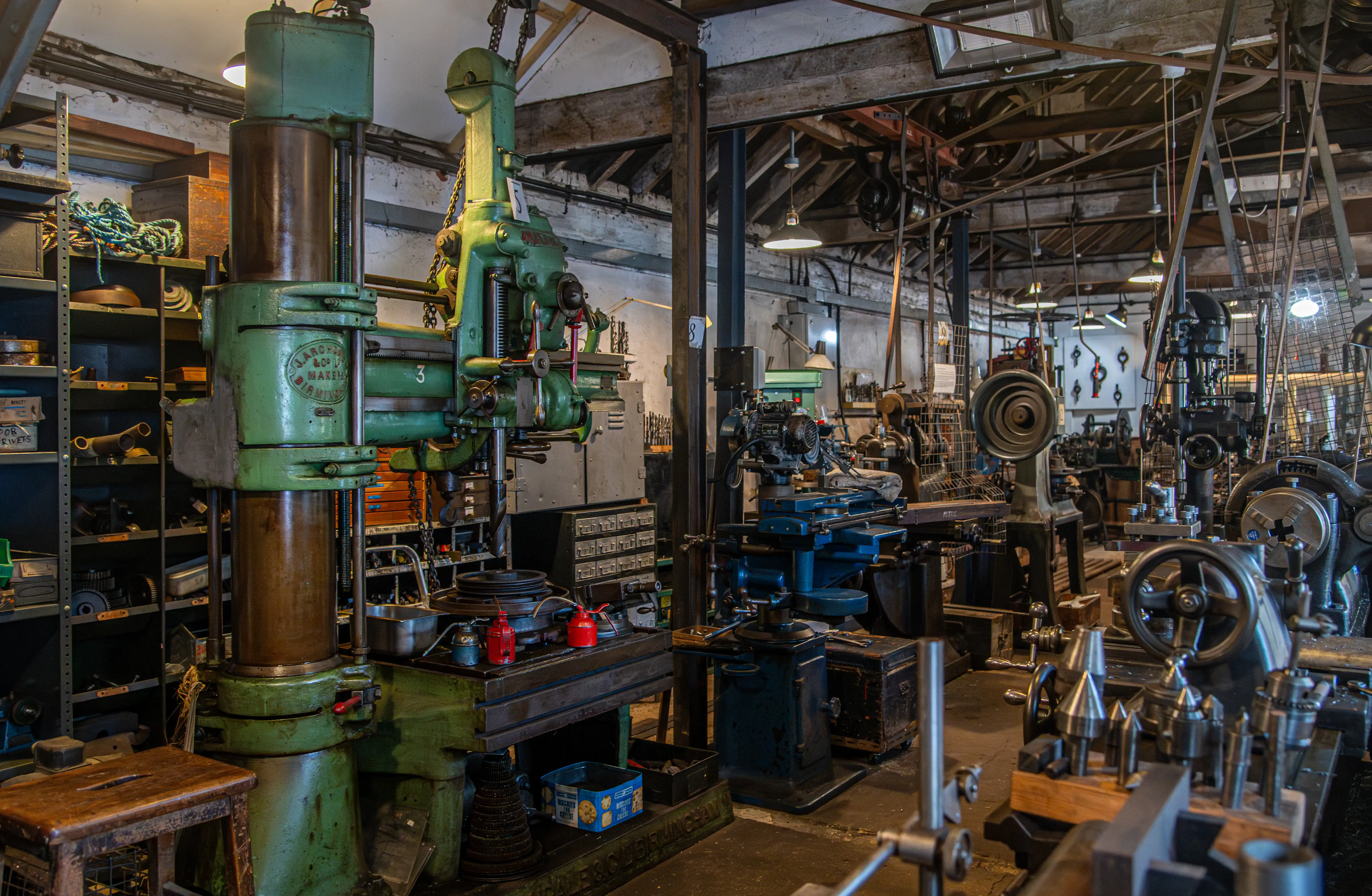 Green milling machine and radial drill press in a traditional workshop with shelves of tools and metal shavings on the workbench.