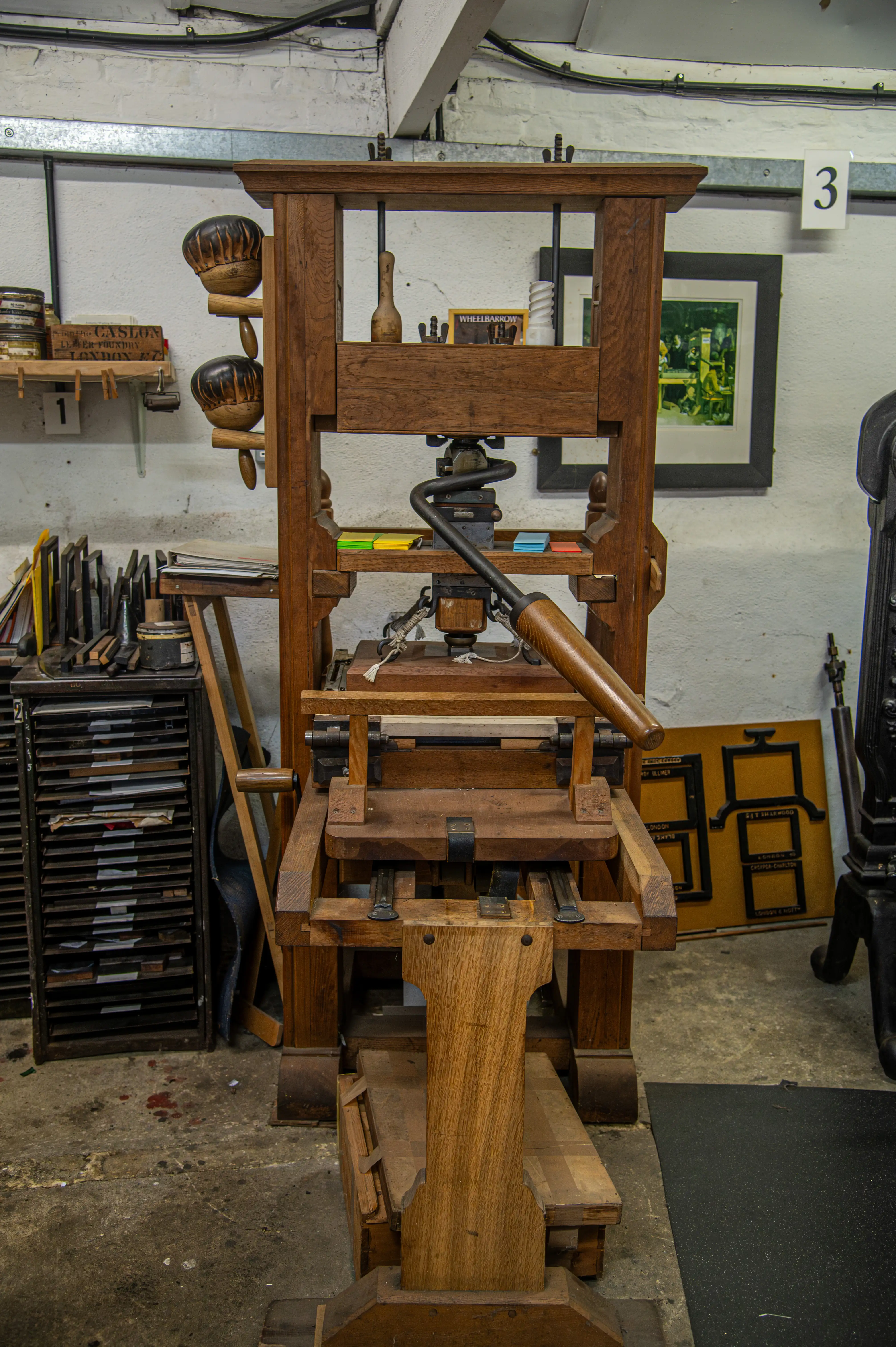 Wooden printing press with screw mechanism, long handle, type trays, ink bowls, and framed prints in a traditional workshop.