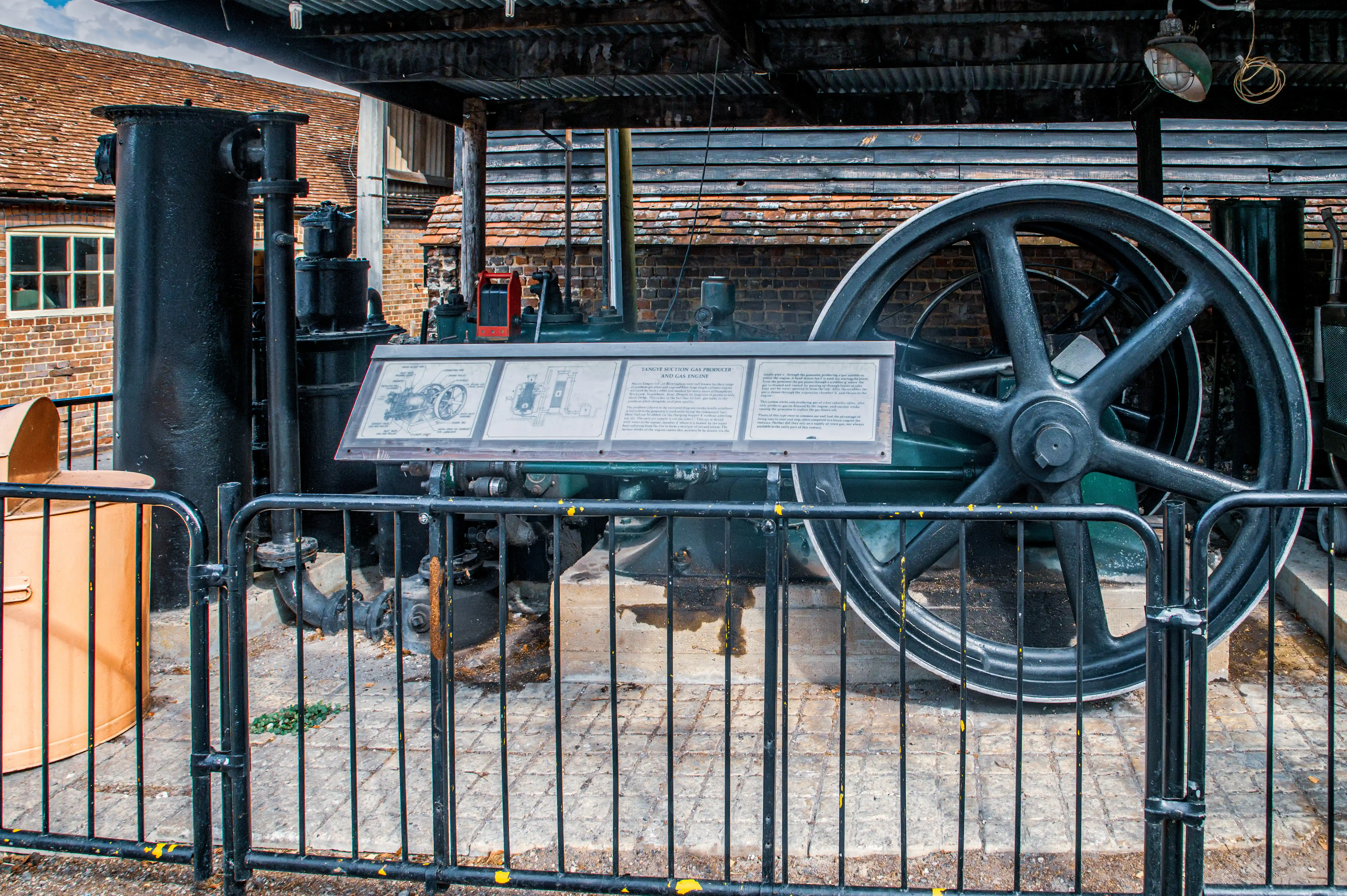 Large stationary steam engine with flywheel and pistons displayed outdoors under a corrugated roof with safety railing and information panel.