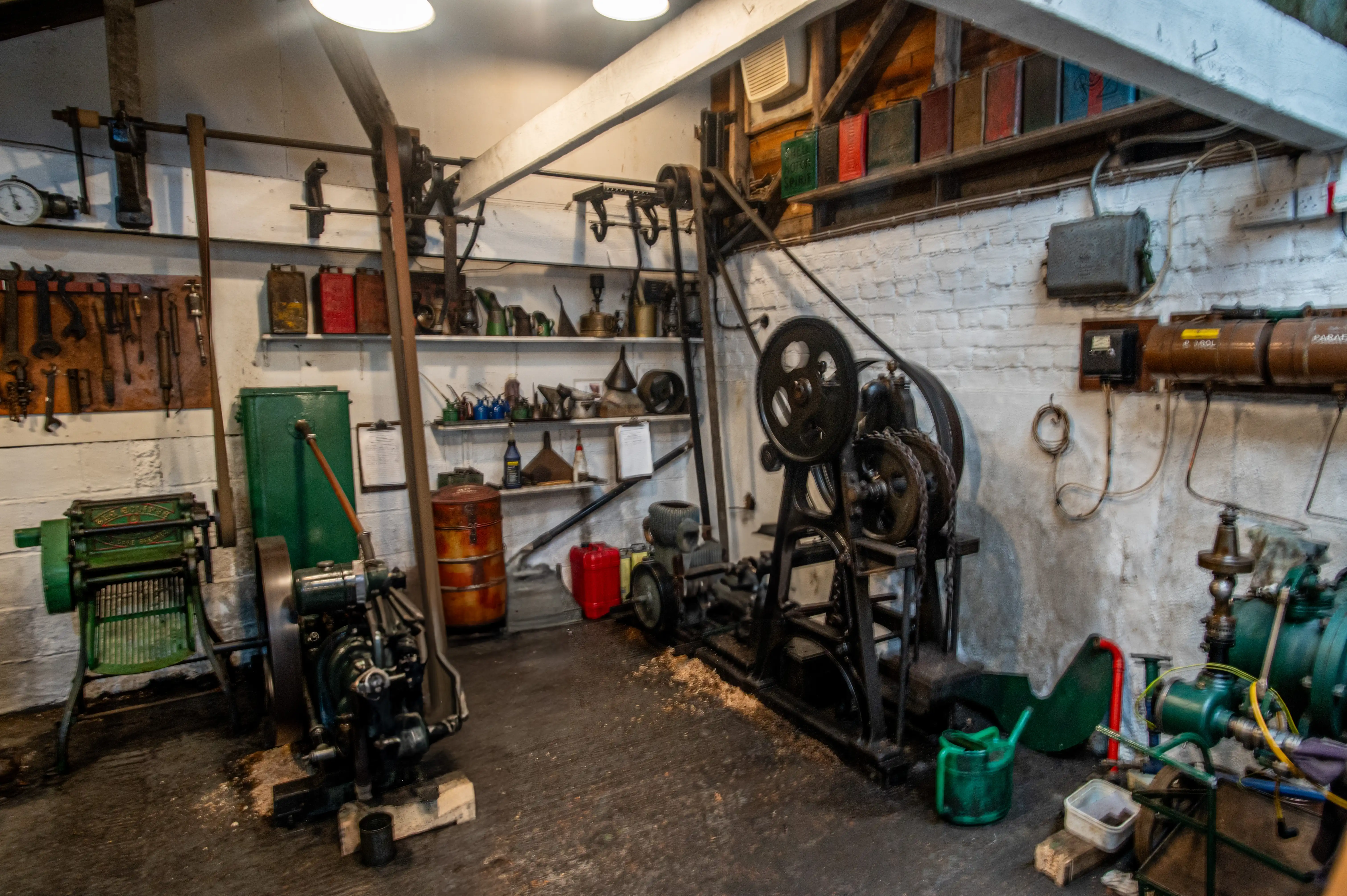 Historic workshop with belt-driven machinery, flywheels, wall-mounted tools, oil cans, and white brick walls.