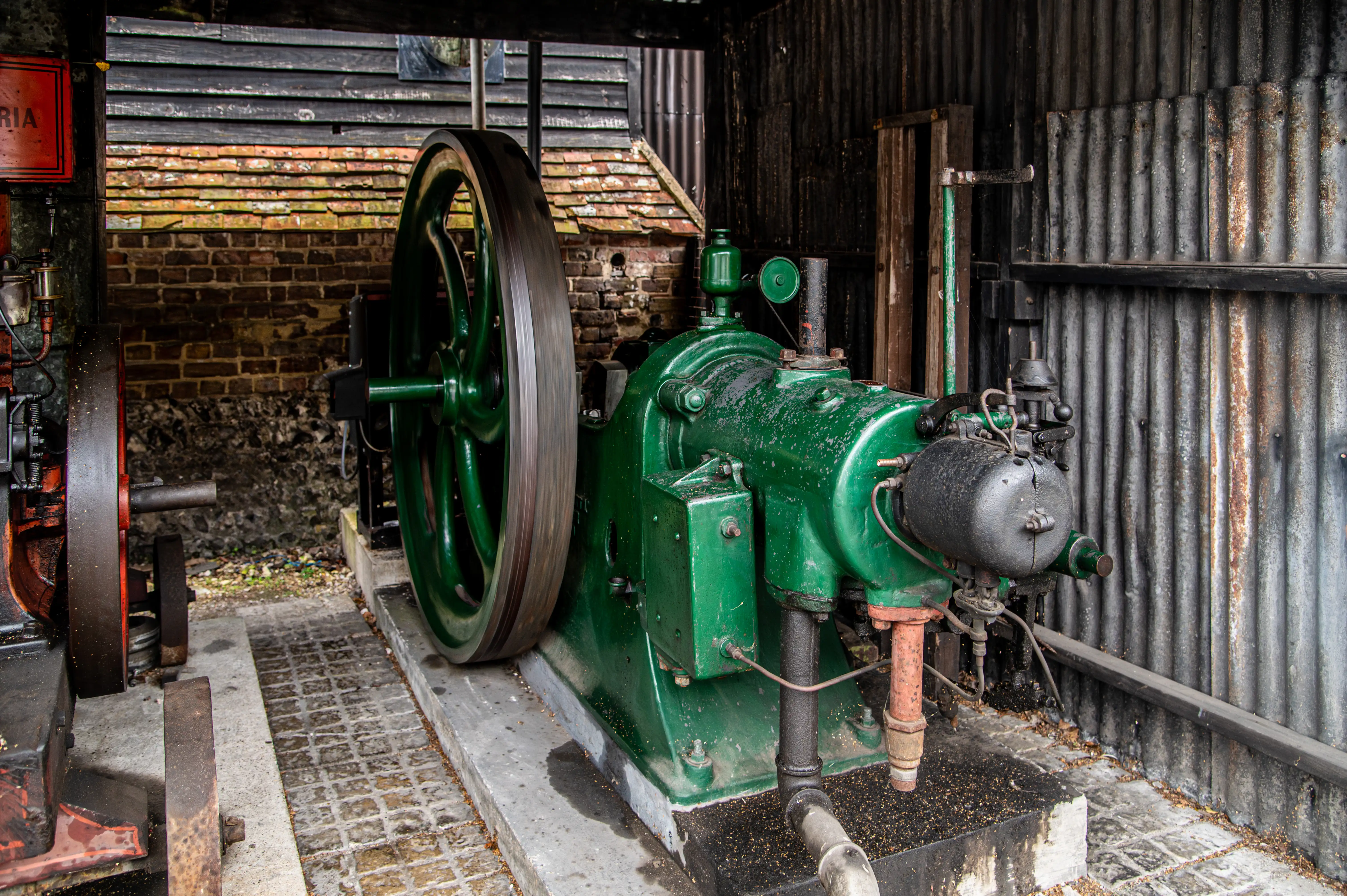 Green vintage stationary engine with flywheel and pipes inside a corrugated-metal shed with brick back wall.