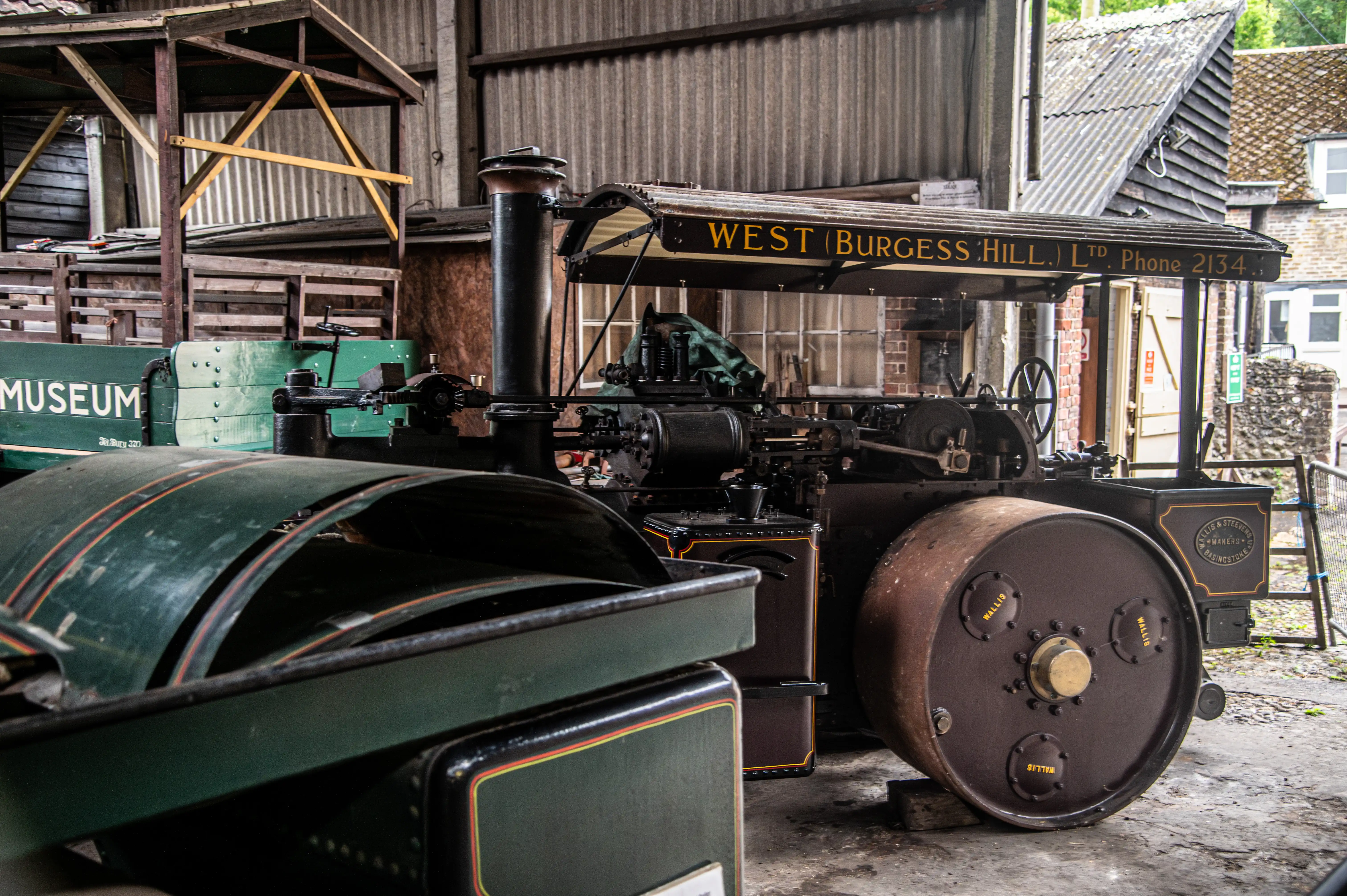Dark-green steam road roller with gold trim displayed in a workshop with wooden structures and corrugated metal walls.