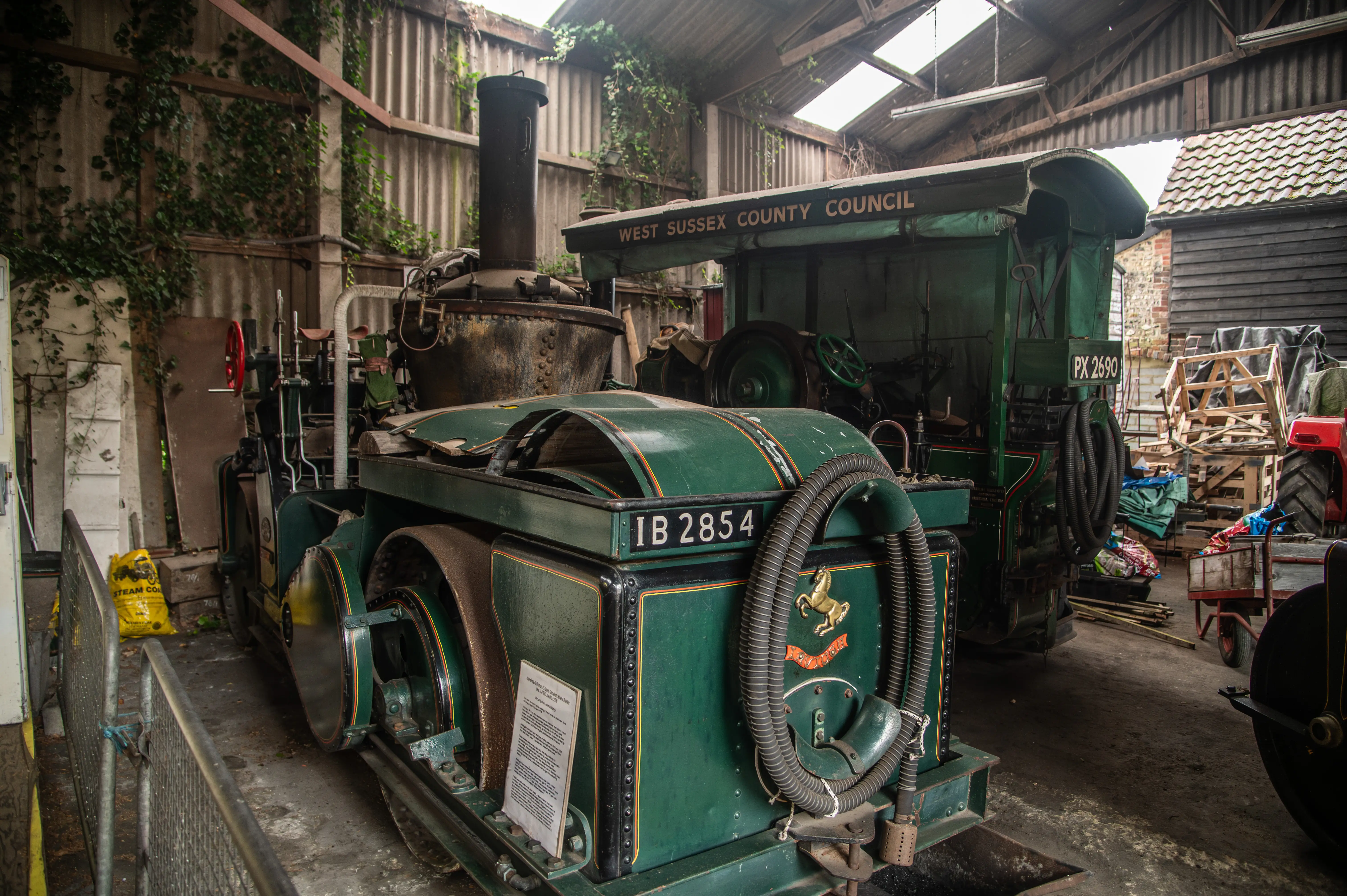Two vintage green steam road rollers with gold and red detailing inside a corrugated shed with ivy, skylights, and surrounding machinery.