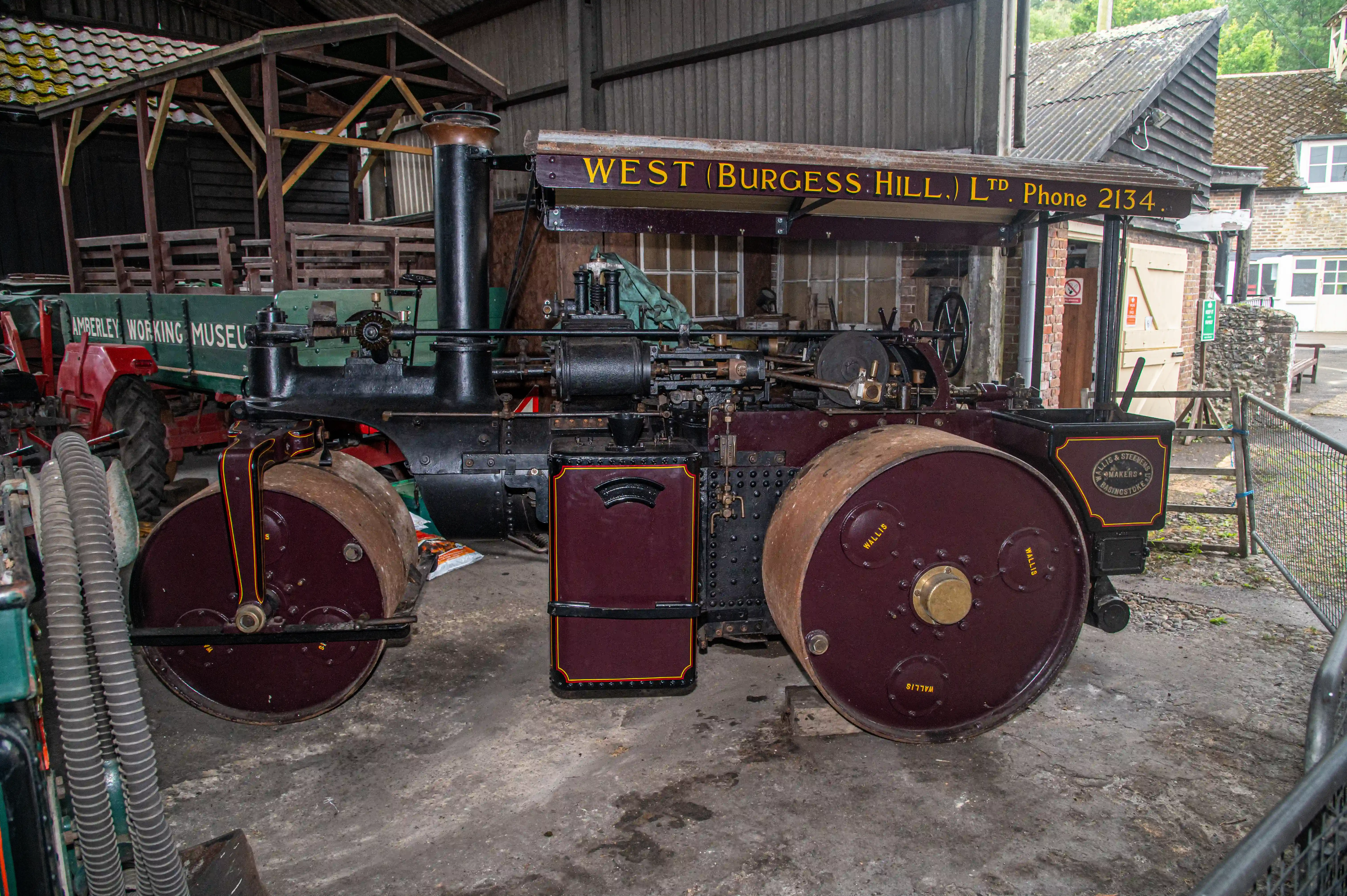 Maroon and black steam road roller with gold trim and Aveling & Porter badge displayed in a museum workshop with a green wagon behind it.